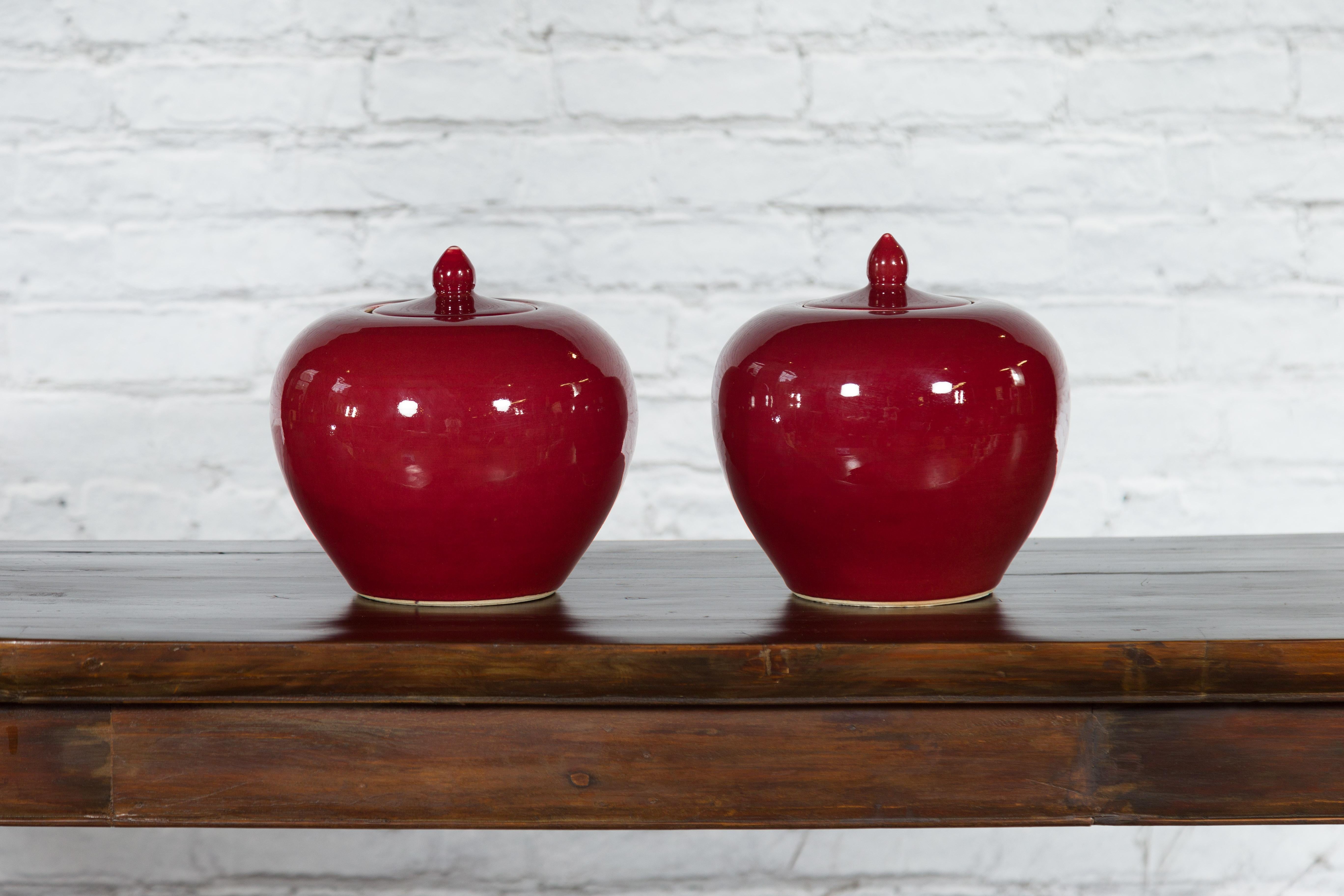 A pair of vintage Chinese jars from the mid 20th century, with oxblood color and lids. Created in China during the Midcentury period, each of this pair of jars features a sleek tapering body perfectly complimented by an oxblood finish. Showcasing