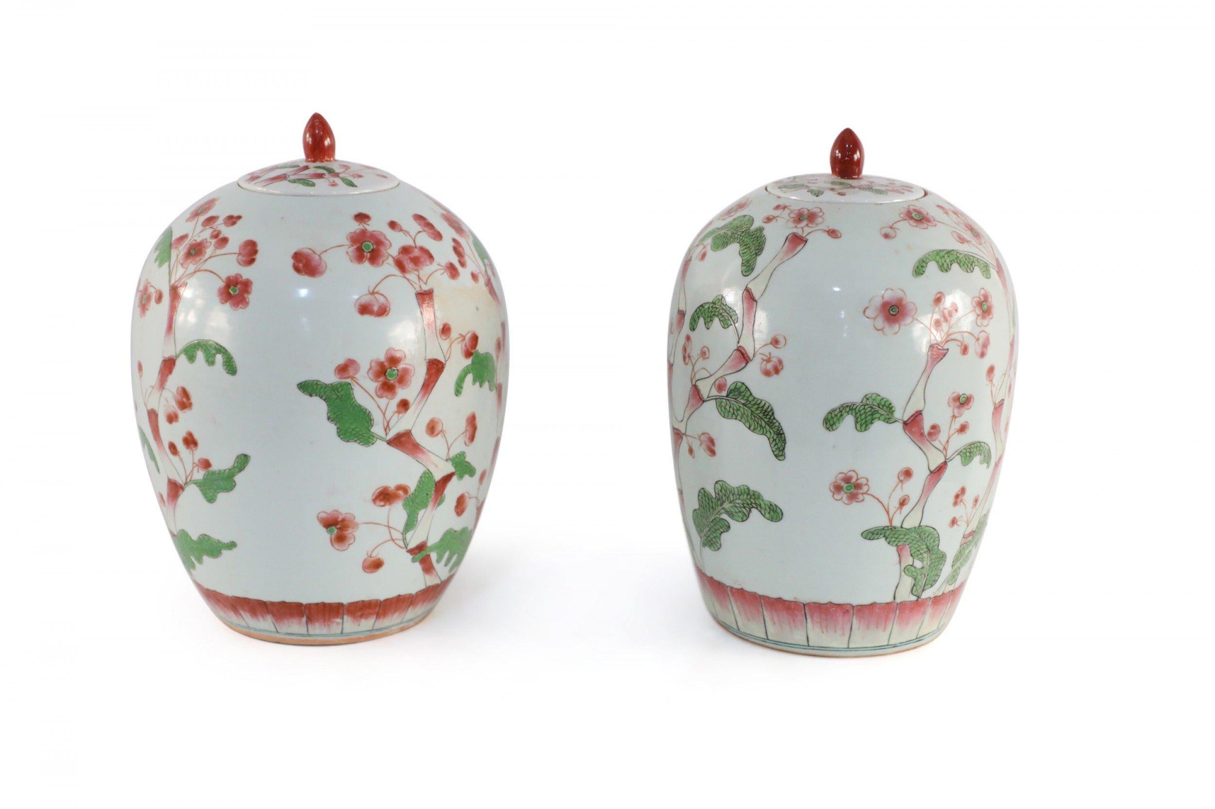Pair of Chinese white porcelain urns decorated with dark pink and green-leafed cherry blossoms growing up the rounded forms, finished with red-finial topped lids and made with holes in the bases (priced as pair).
      