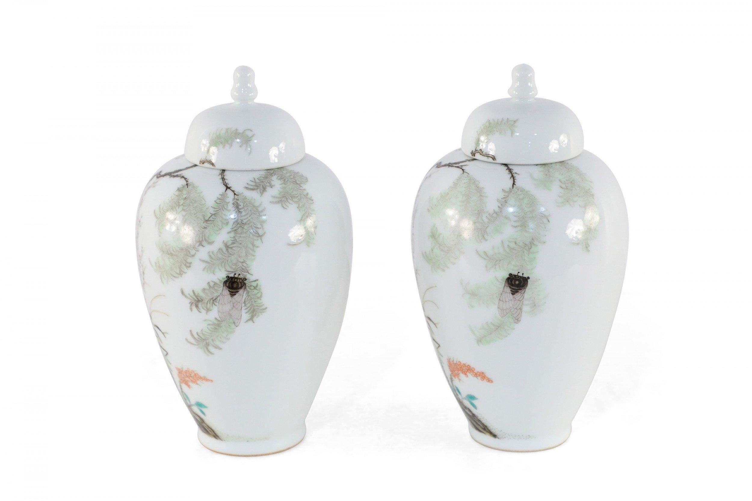 Pair of Chinese white porcelain famille rose jars painted with verdant trees and insects with characters on the backs, finished with finial-topped lids.