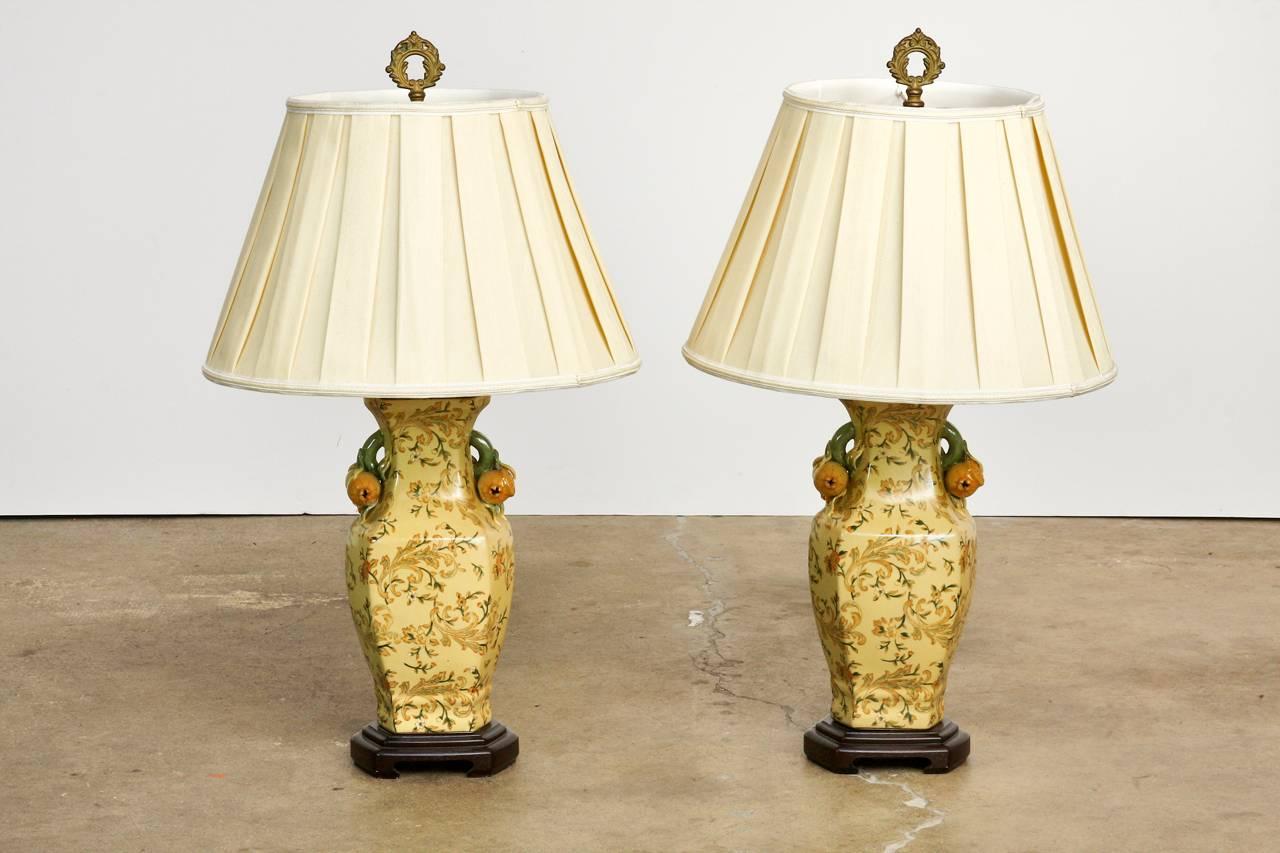 Remarkable pair of chinoiserie porcelain vase table lamps by Bradburn Gallery. Each lamp features a floral and scrolling vine design with porcelain peach handles on each side. Mounted to a wooden base with an Asian motif and topped with polished