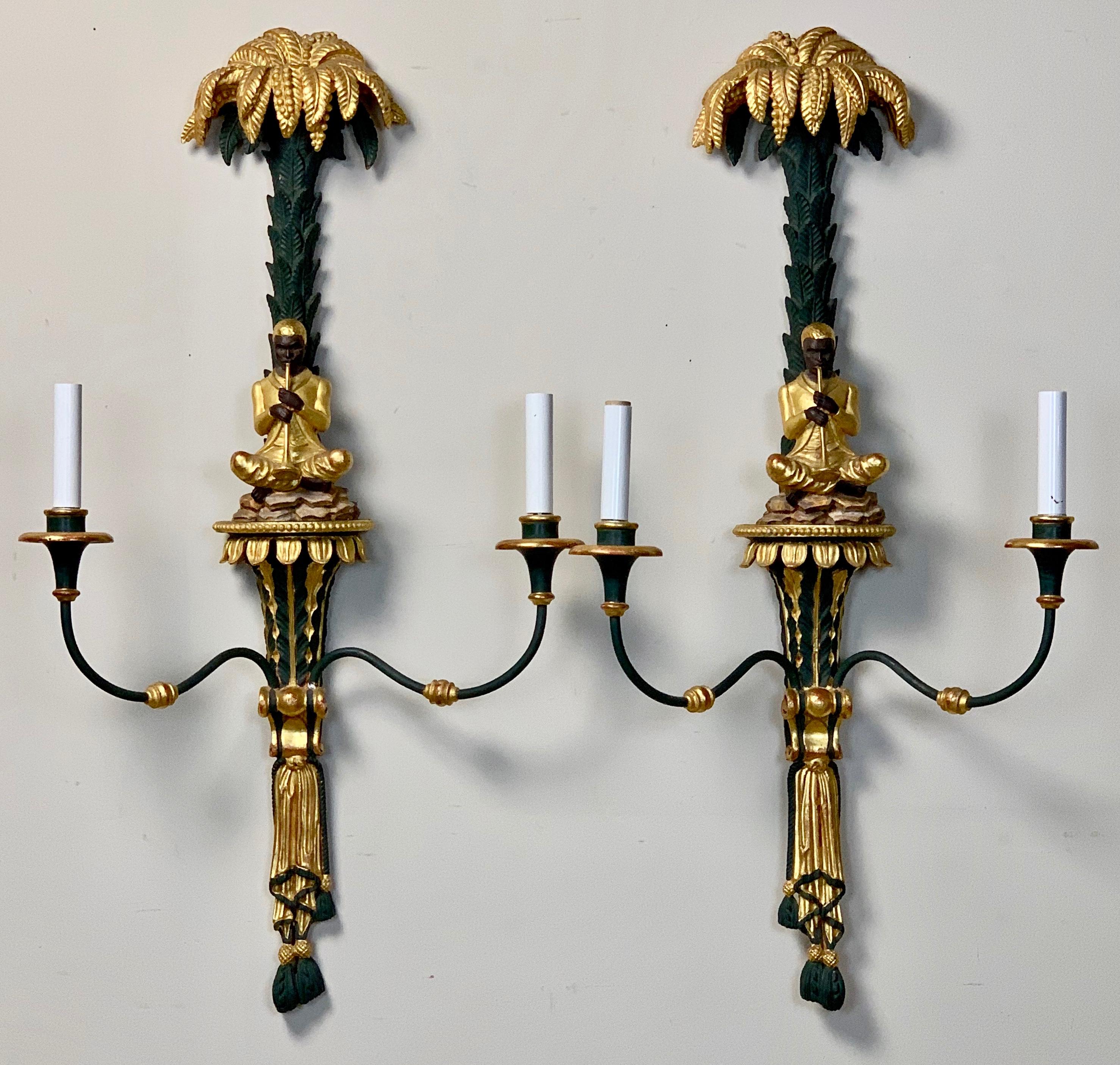 A whimsical pair of mid-20th century Regency style carved wood deep green painted and water gilt decorated electrified sconces depicting seated flute players under a palm trees.