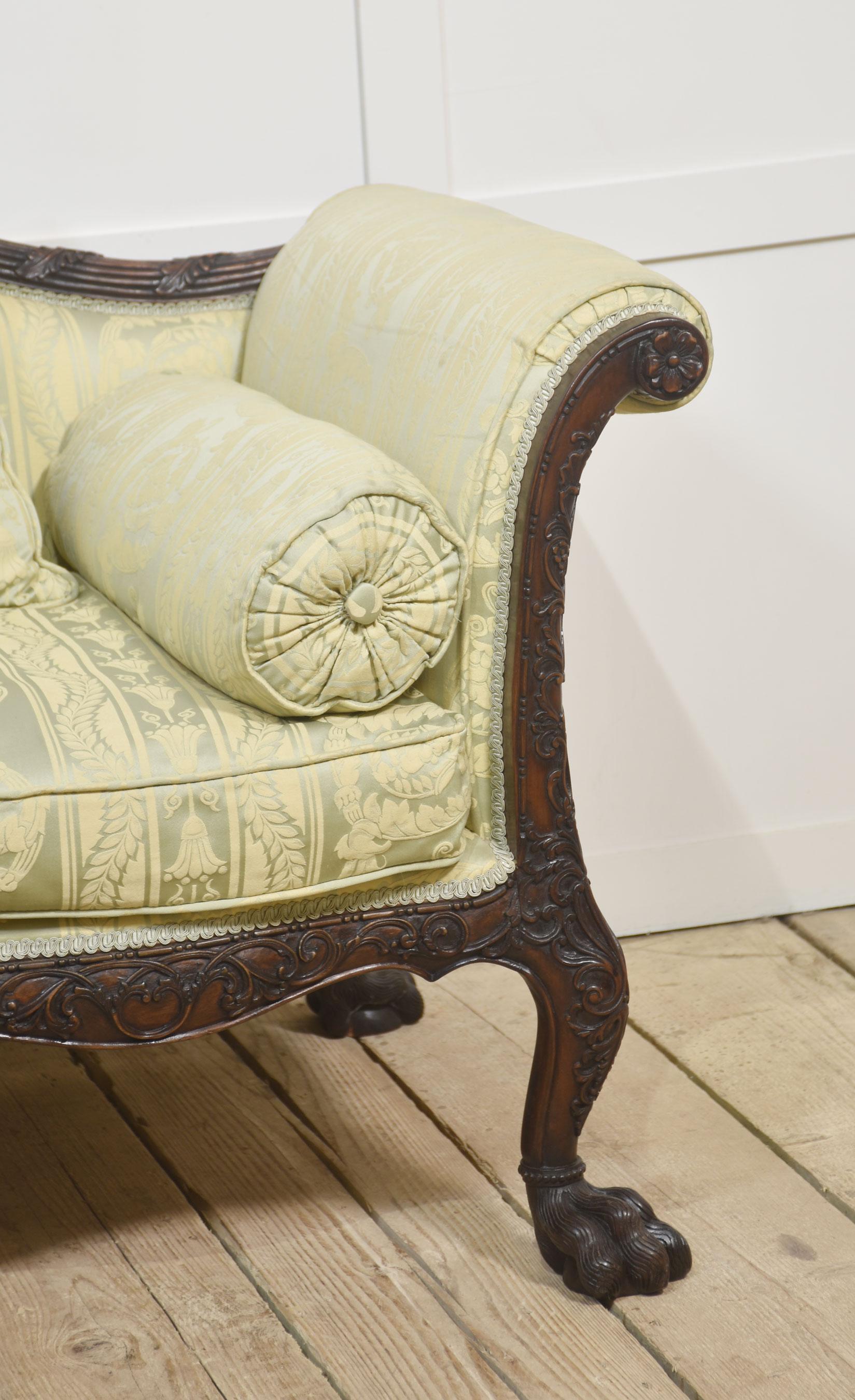 A striking pair of settees featuring a finely carved mahogany frame with intricate detailing along the crest rail and apron. The high, arched back is complemented by elegant, scrolled arms and rests on bold, claw-and-ball feet, showcasing masterful