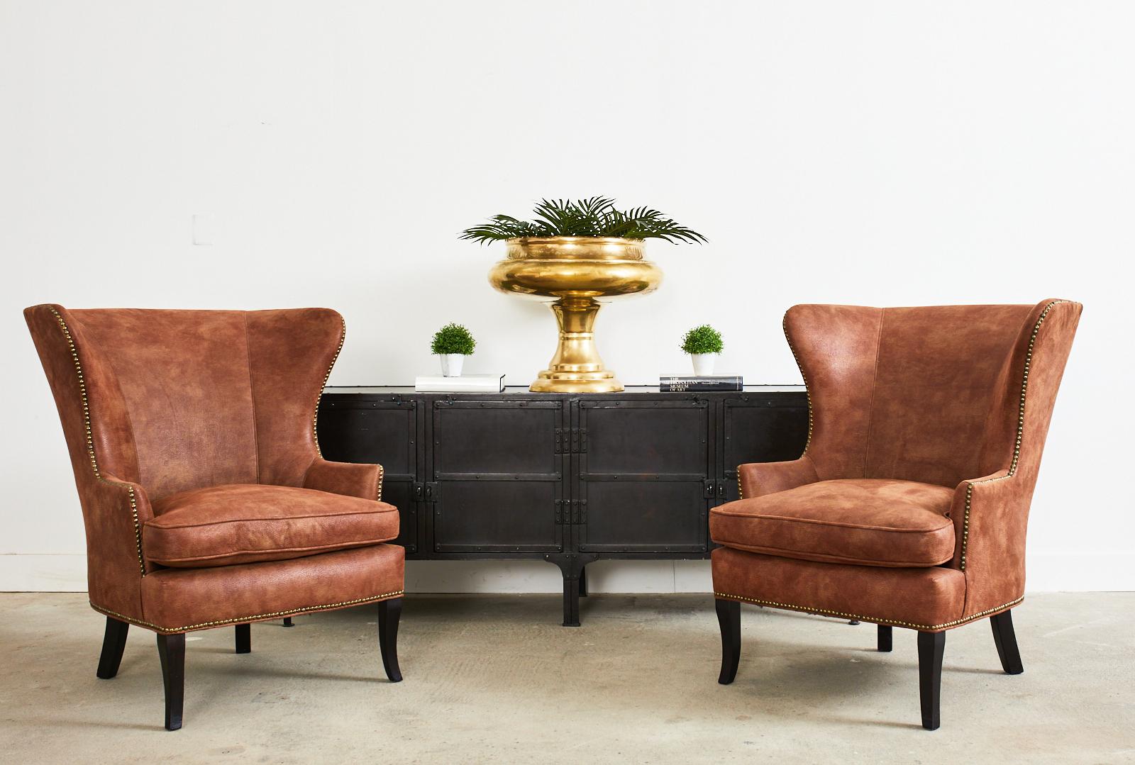 Distinctive pair of modern wingback armchairs featuring an intentionally distressed faux-leather upholstery by Christian Audigier. Crafted from hardwood frames with large, wide wings gracefully curving down to the narrow arms. Bordered by brass tack