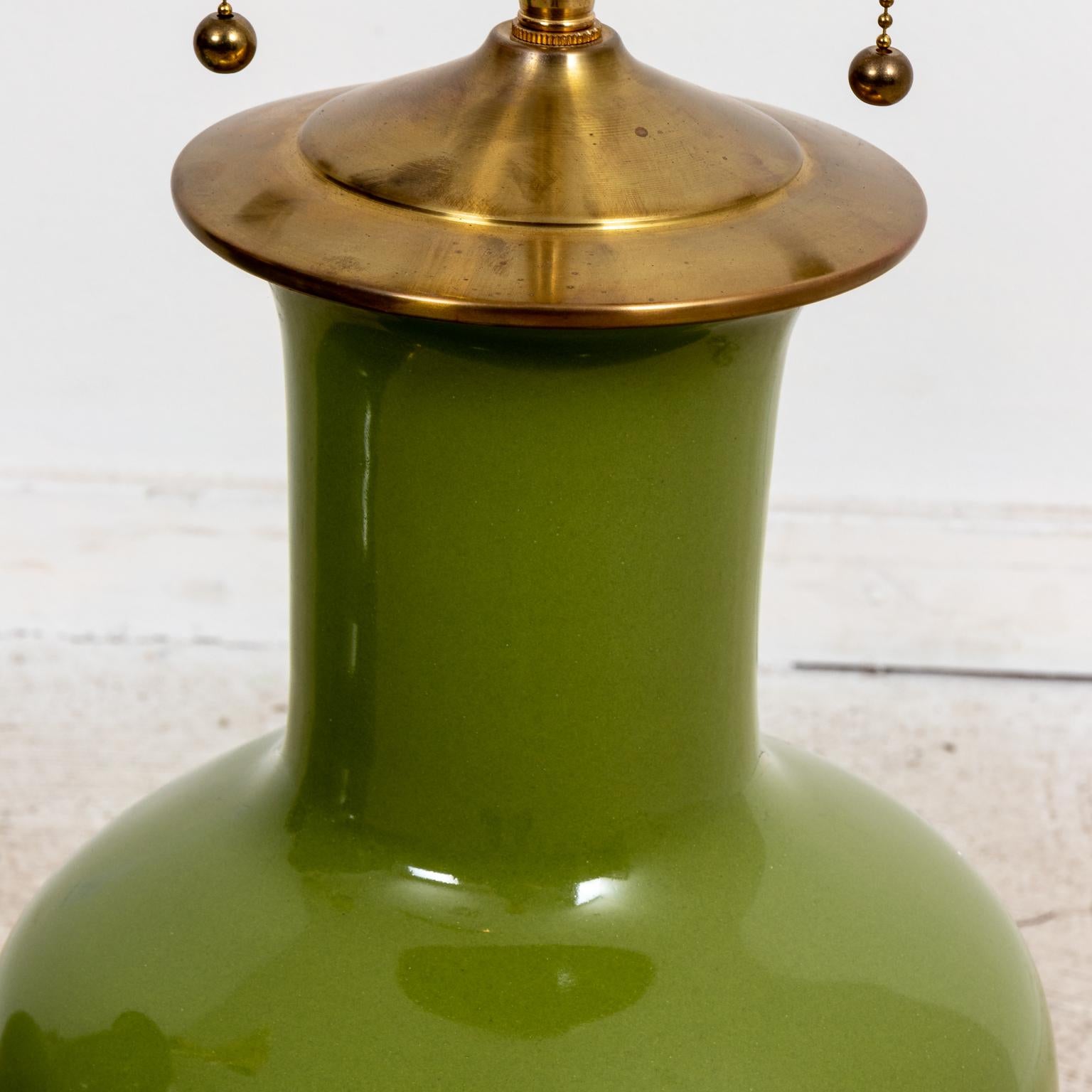 Circa 21st century Contemporary style large pair of Avocado Green ceramic table lamps by Christopher Spitzmiller with 14 karat gold gilt bases. The lamps are further constructed with brass necks, double light sockets, and ball finials. Made in the