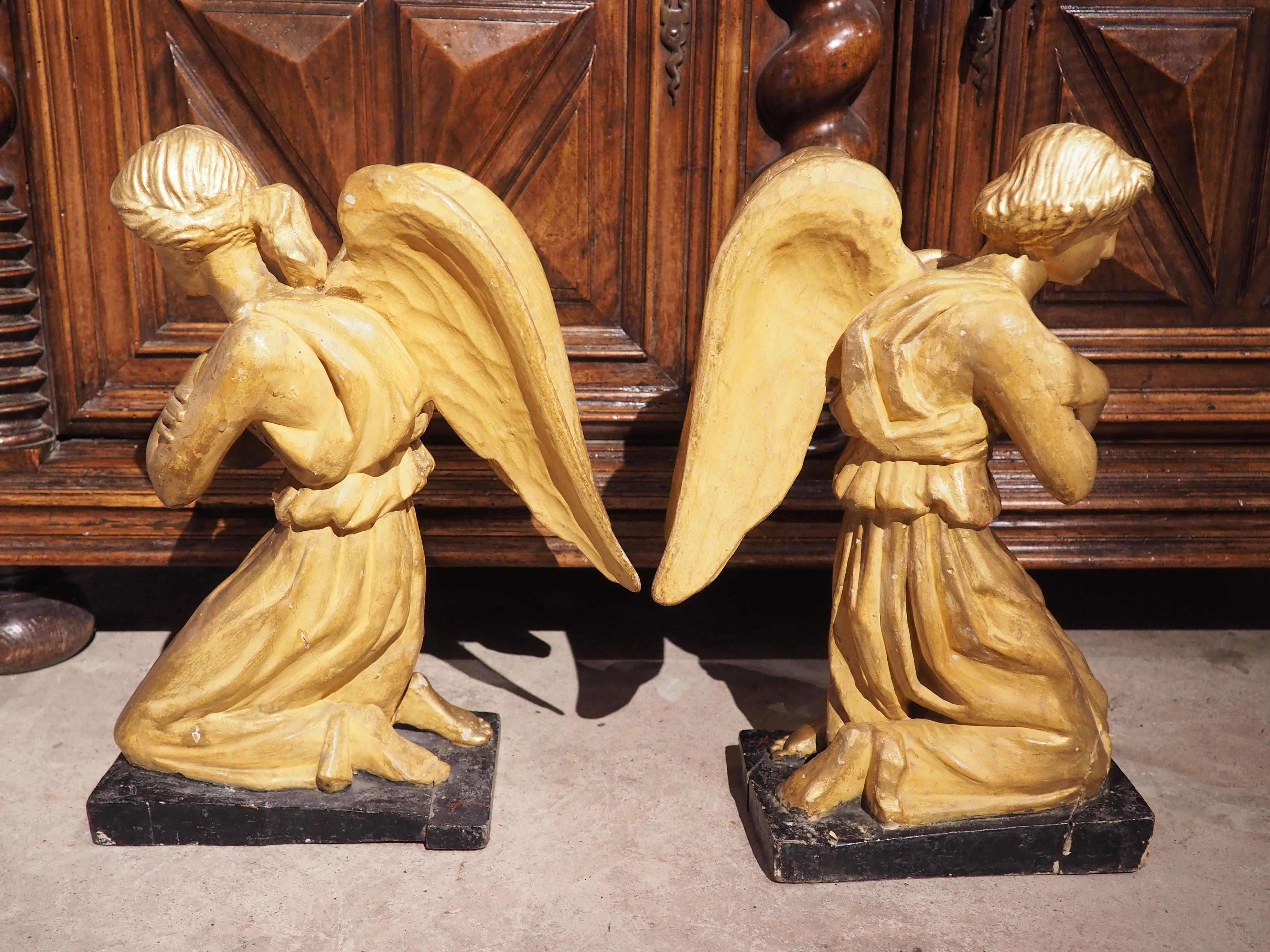 Hand-carved in Italy, circa 1800, this pair of giltwood angels is depicted in a double genuflection, or kneeling, stance. Both angels are completely gilded with addorsed wings and a long, flowing chiton gathered by a brooch at each shoulder. The