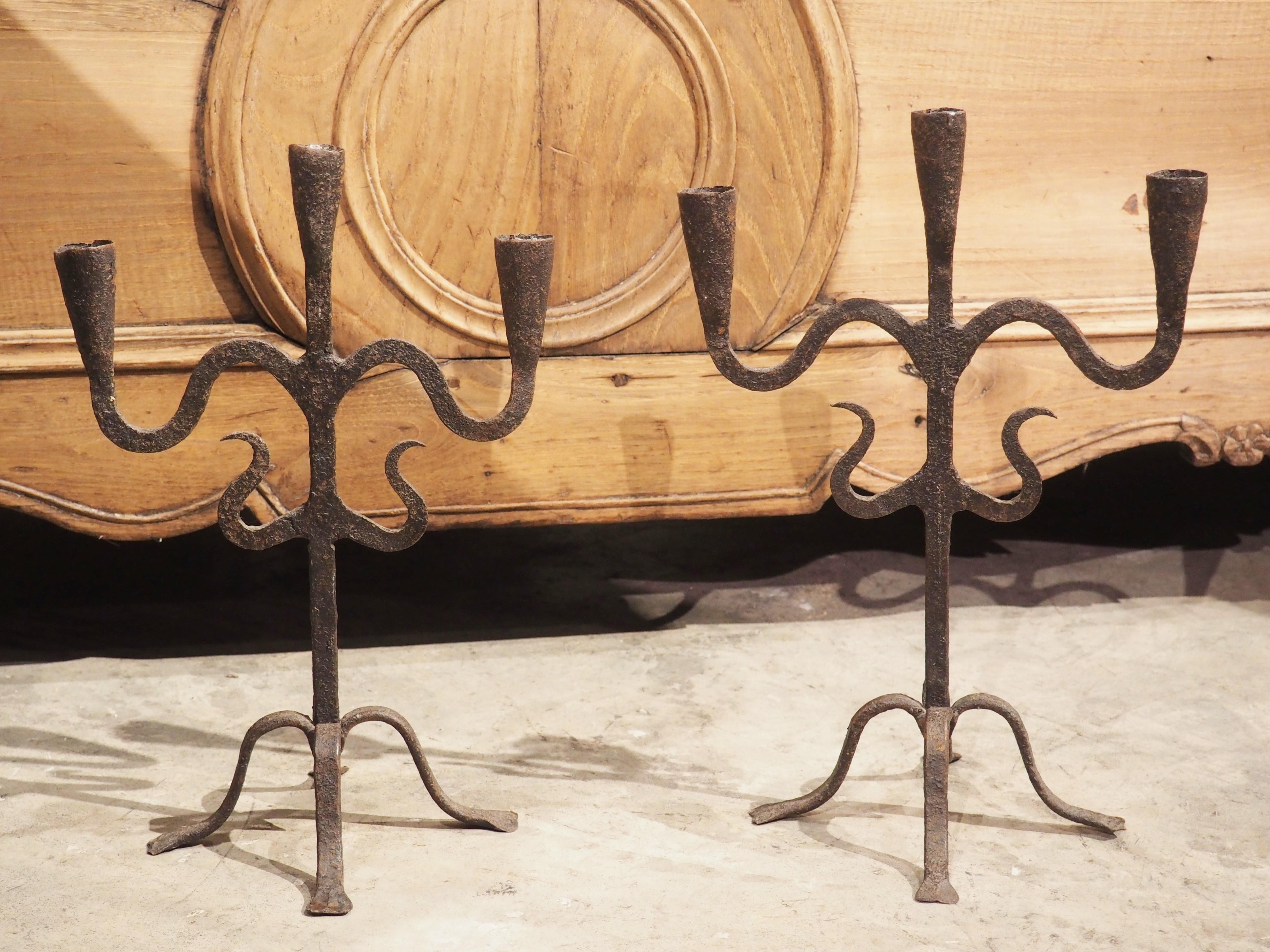 Hand forged in Spain, circa 1800, this pair of iron candle holders can be used in any room of the house. Both candle holders have three cylindrical cups, with the center one sitting above the outer two that rest on S-scroll arms. A pair of smaller