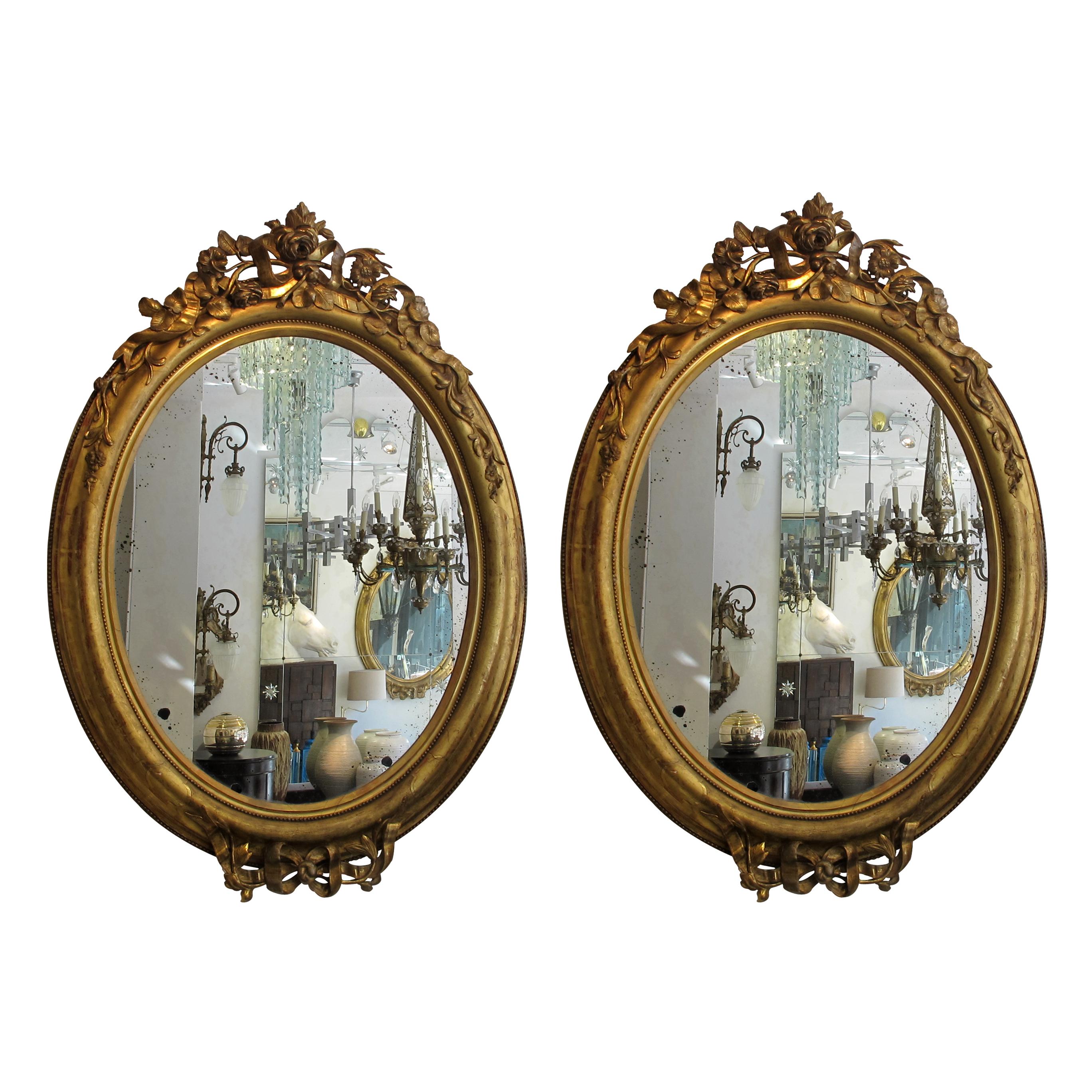 A pair of large French Napoleon lll oval mirrors with giltwood frames and original glass, circa 1860.
The crest holds elaborated carved wood roses and ribbons, the bottom of the oval holds a very elegant ribbon tie with a flower bud. The frame is