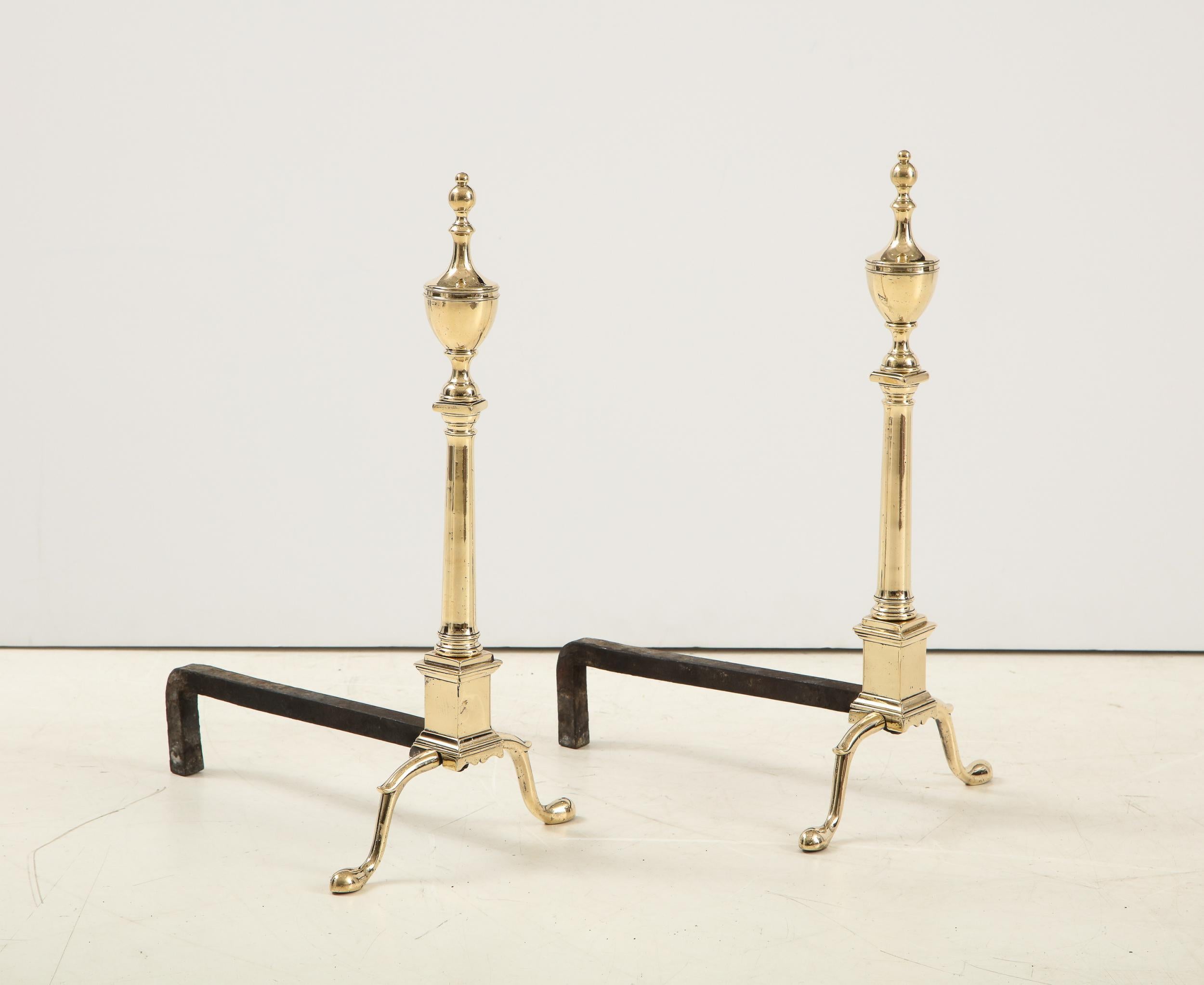 Fine pair of American classical Federal andirons, having urn finials over Doric column shafts standing on square plinths with scalloped base, over shaped legs having spurred knees and cabriole legs. The metal is either a pale bell metal or brass