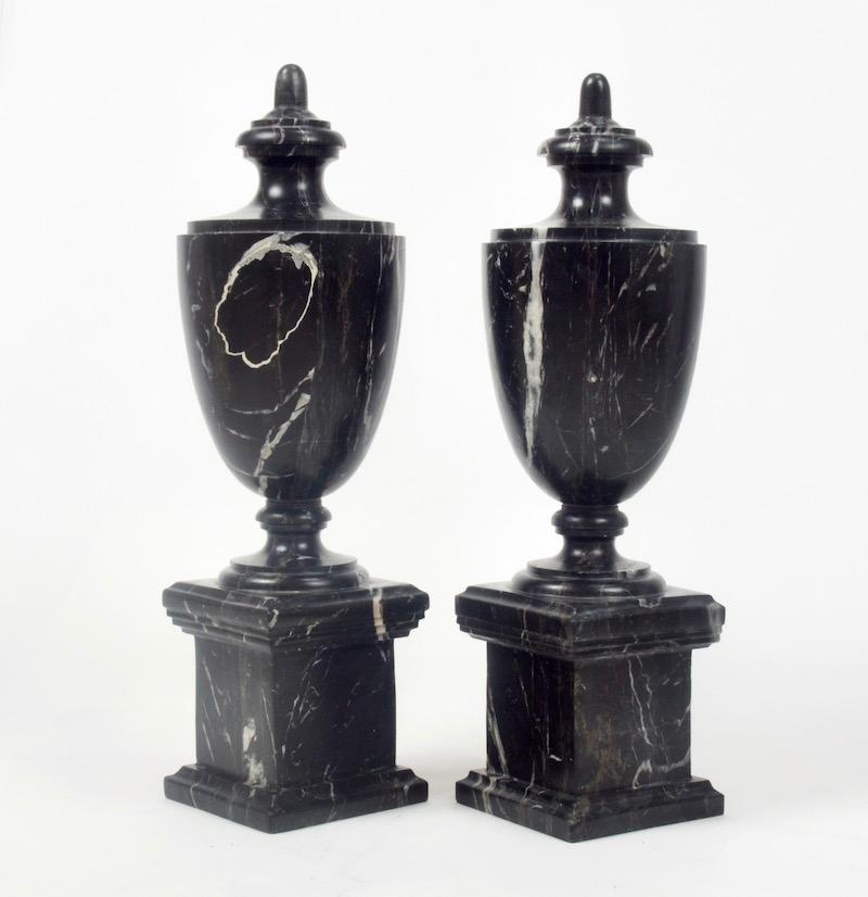 A pair of decorative early 20th century black marble urns of classical shape set upon pedestals surmounted with faux-lids. Carved from a single piece of stone the rich deep black marble is contrasted by beautiful dappled white crystalline streaks.