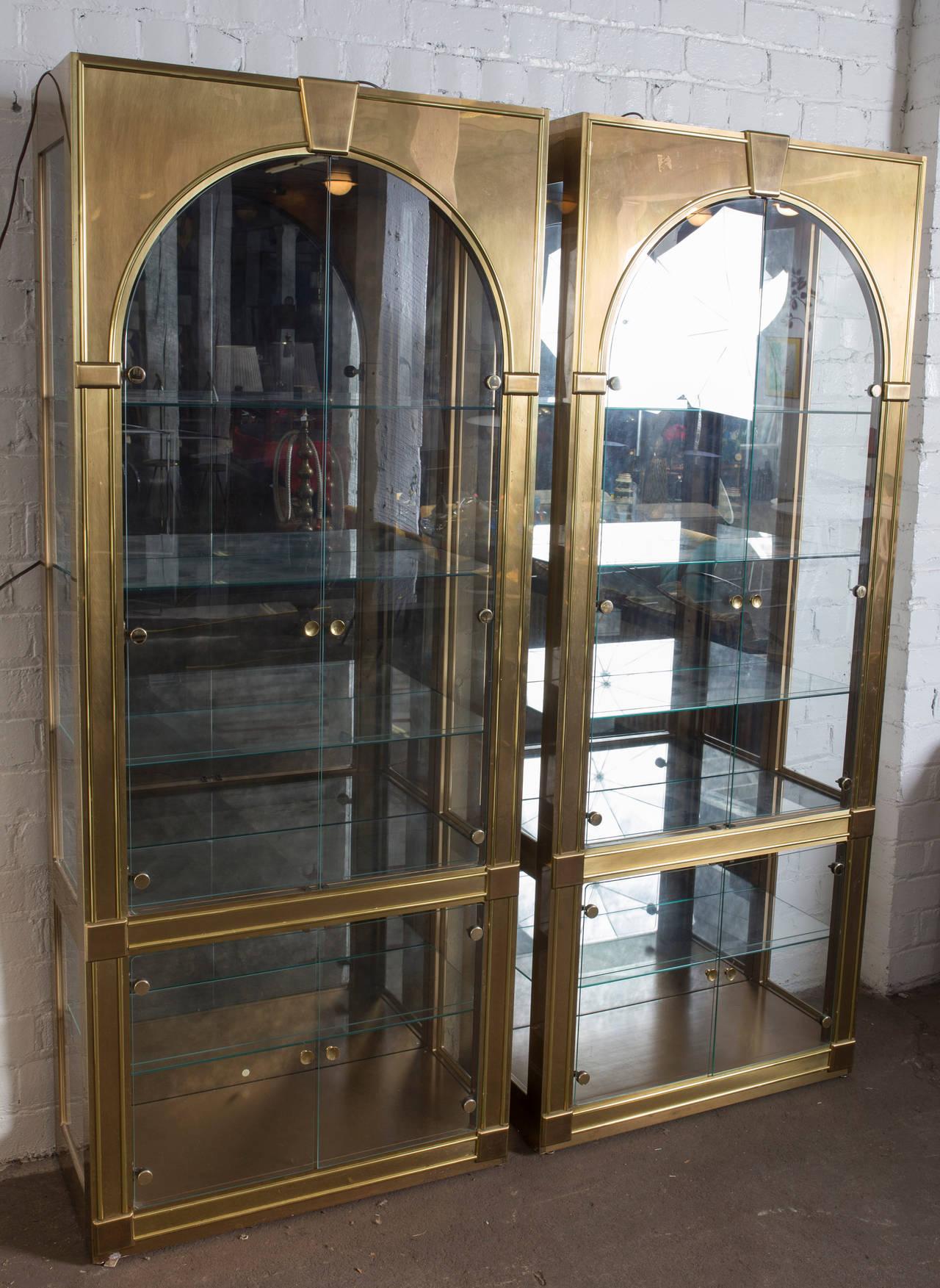A pair of impressive vitrines in the classical Palladian style, manufactured by Mastercraft, circa 1970s. Each with two-door arched fronts, antiqued smoked mirrors backs, clear glass against brass frames. The interior with three glass shelves above