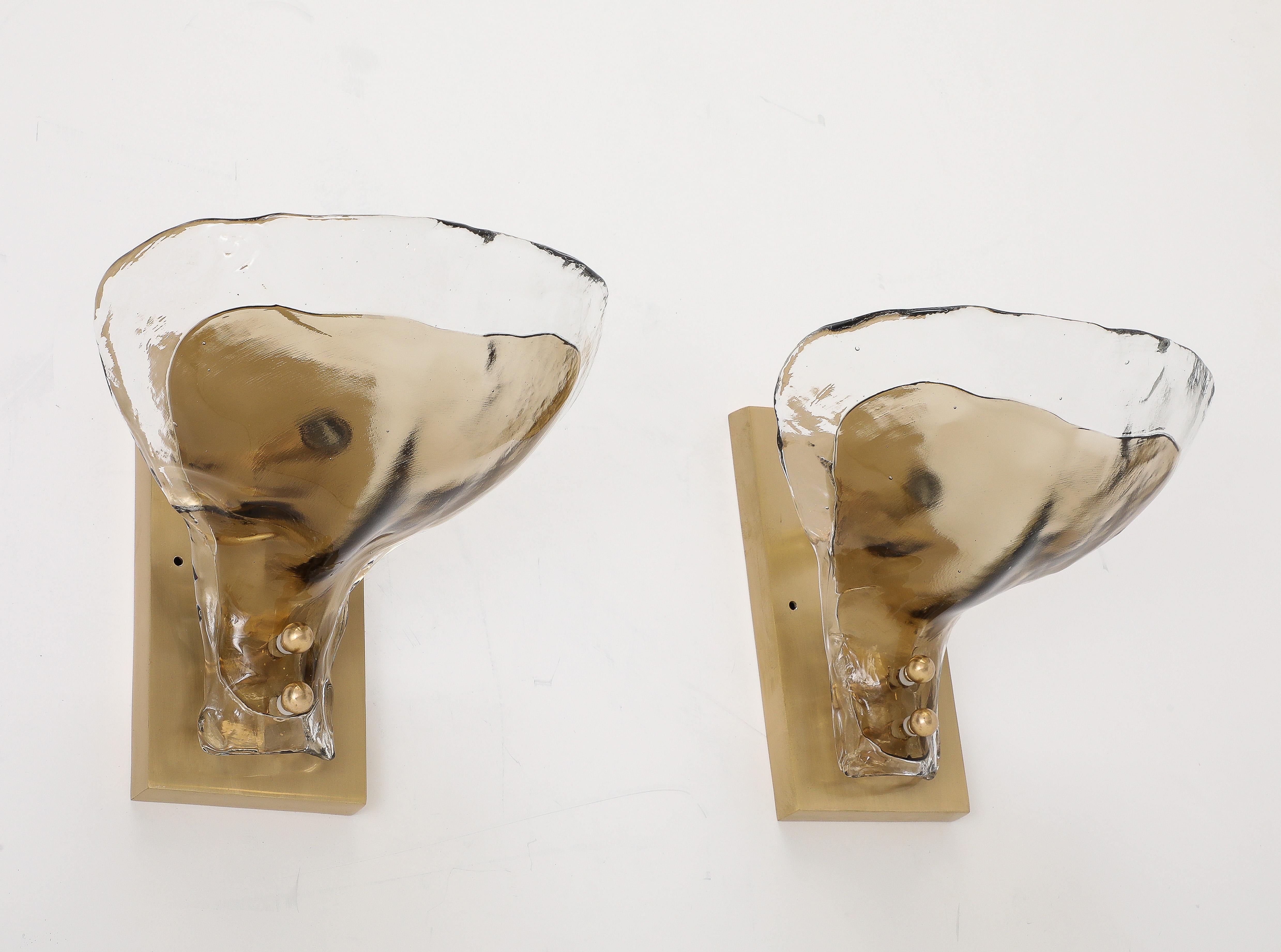 Coppia di appliques minimaliste in vetro di Murano trasparente e tabacco di Mazzega.
Lo spesso petalo a forma di foglia in vetro di Murano è montato su una piastra in ottone lucido ed è dotato di
una singola presa a candelabro che è stata appena