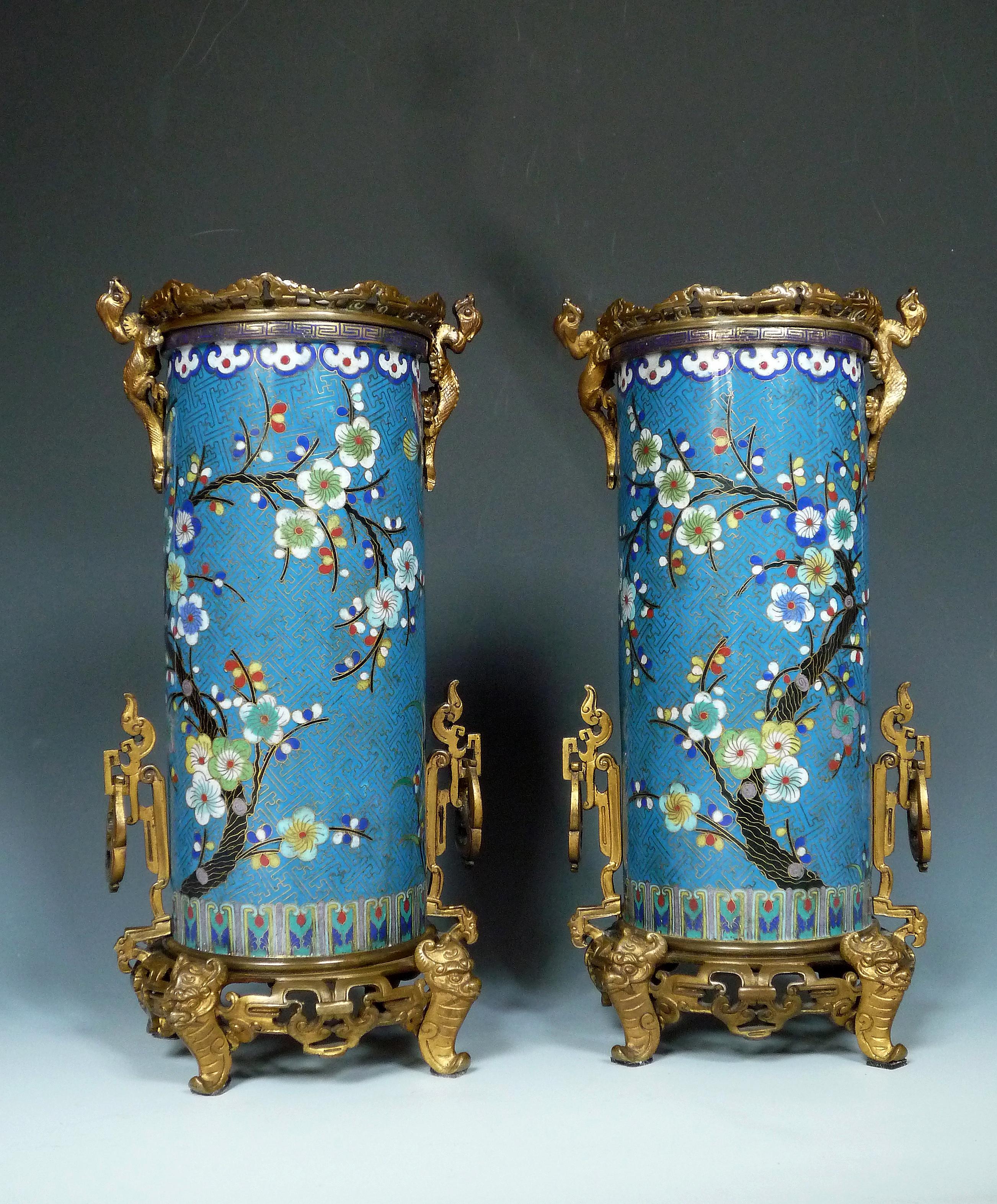 Pair of Japanese-style roll-shaped vases, made in “cloisonné” enamel and gilded bronze. The cylindrical body is decorated with polychrom floral branches, flowers and butterflies on a blue background. The mounts made in pierced and “old gold”