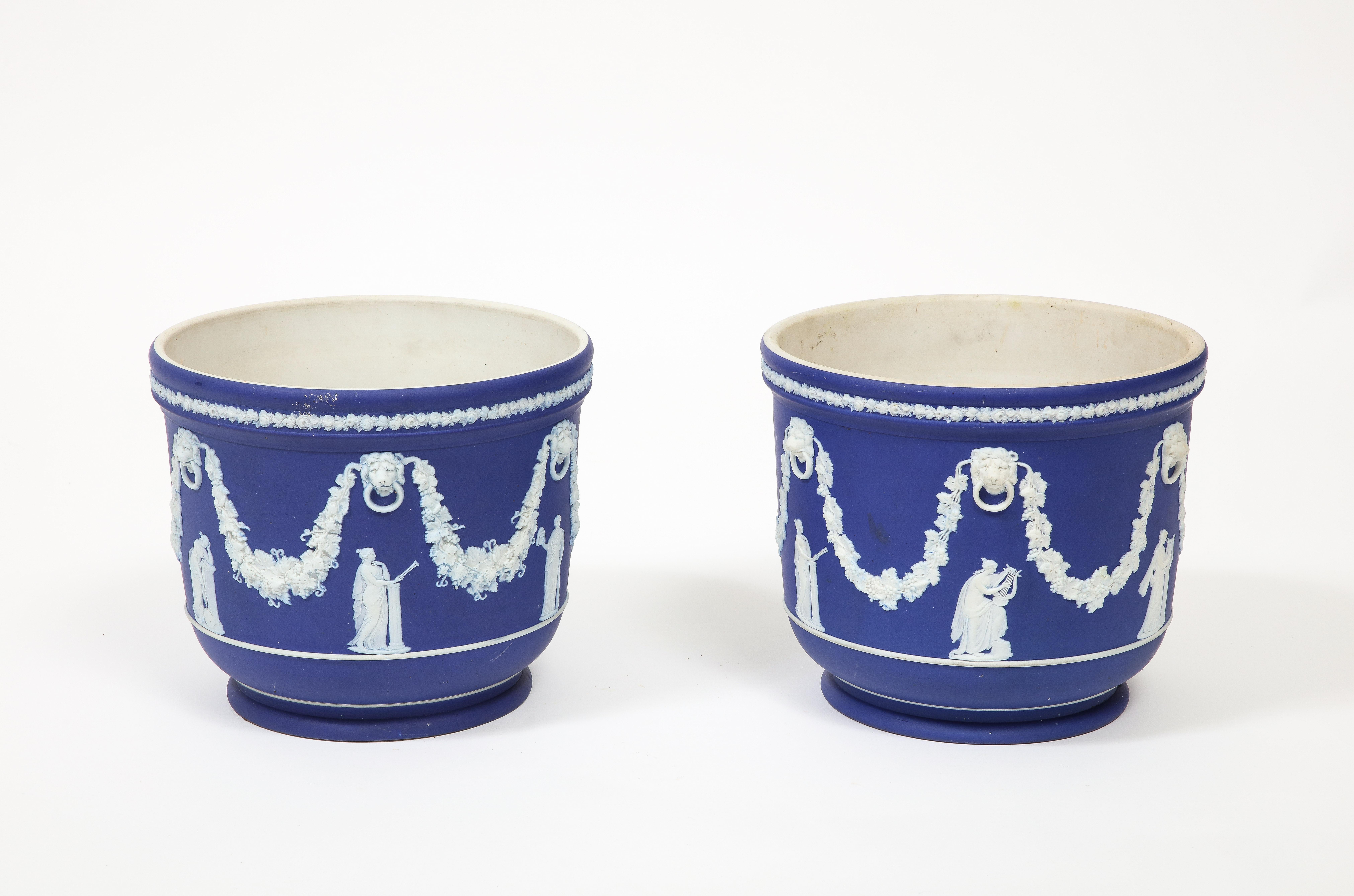 Pair of early 20th Century antique cobalt Wedgwood Jasperware planters, featuring classical figures beneath lion's heads and garlands of grapes. Along the top edge is a band of fruits and flowers. The interior is white porcelain. 