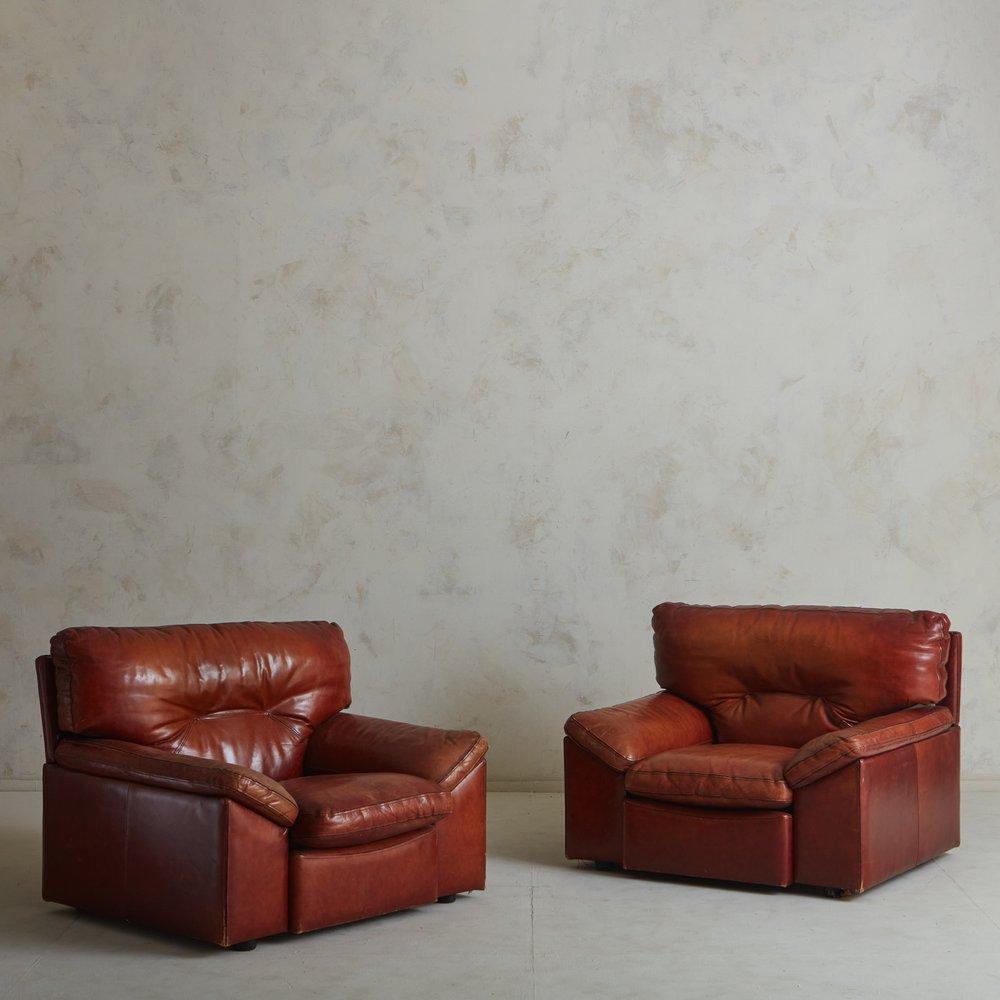 A pair of 1960s French lounge chairs. These chairs retain their original patinated cognac leather and feature stately profiles with angled arms. Each chair has two removable cushions with stitch detailing and is elevated off the ground with circular