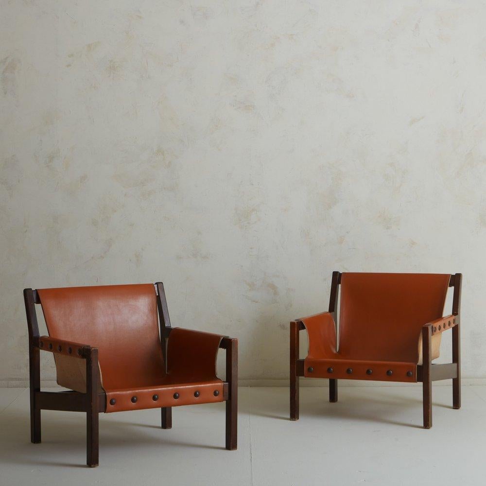 A pair of 1970s French lounge chairs featuring handsome cognac leather slingbacks and seats with stitch detailing. The leather is affixed to the angular wood frames with circular metal studs. Unmarked. Sourced in France, 1970s.