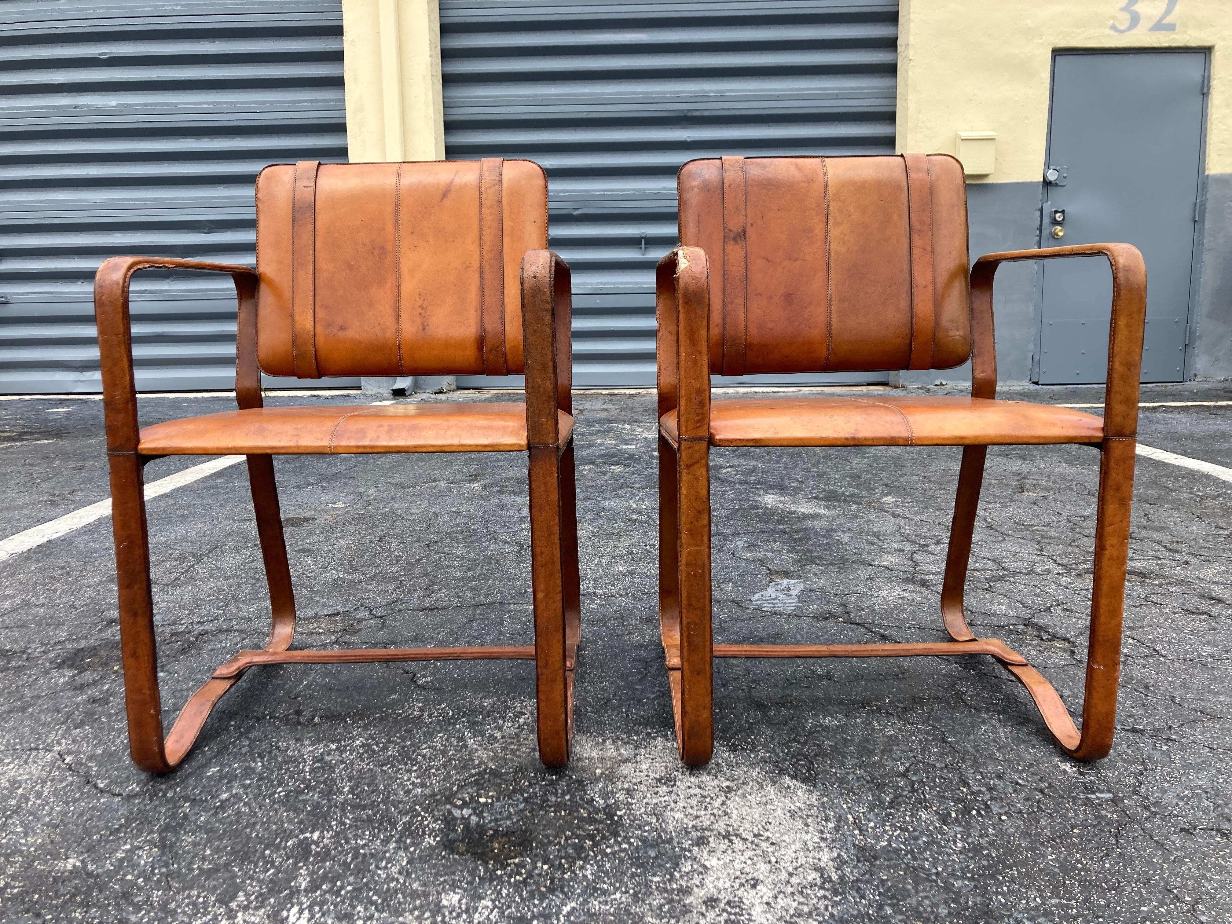 Pair of leather wrapped armchairs in the style of Jacques Adnet, Cognac leather with strong patina and damages. The chairs are sturdy.
Arm height is 27”.