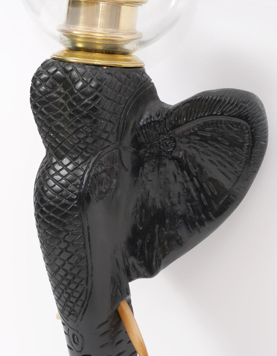 This exotic colonial style pair of candle wall sconces feature carved and ebonized elephant heads with bone tusks holding stylized pineapples in their trunks. On top of the elephants elegant brass mounts hold candle holders and handblown brass