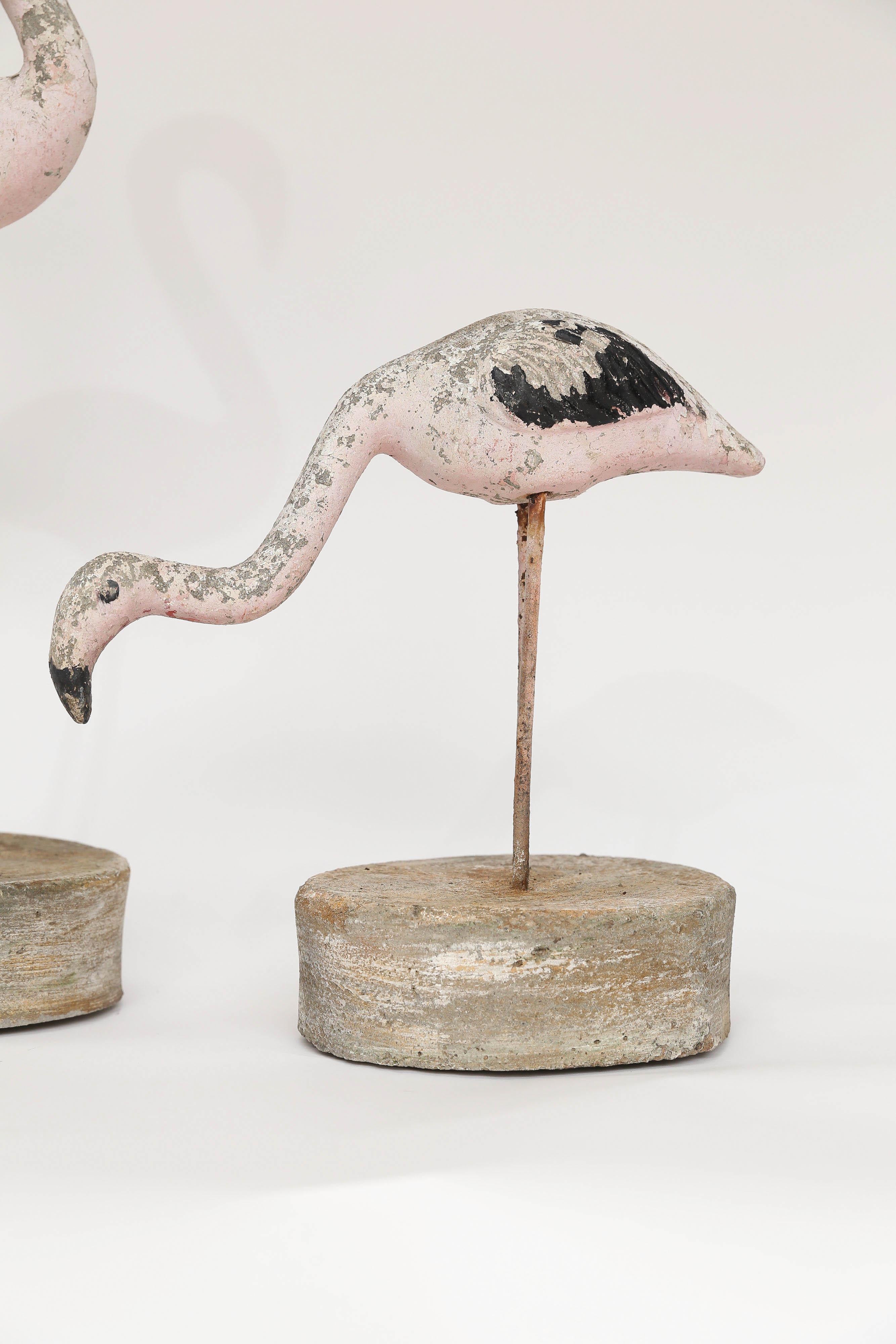 A lovely pair of concrete flamingos. The concrete birds stand on legs made of iron sunk into a hefty concrete base. The flamingos retain original paint which has been worn to a lovely patina. The upright flamingo body measures 7 inches wide and 3