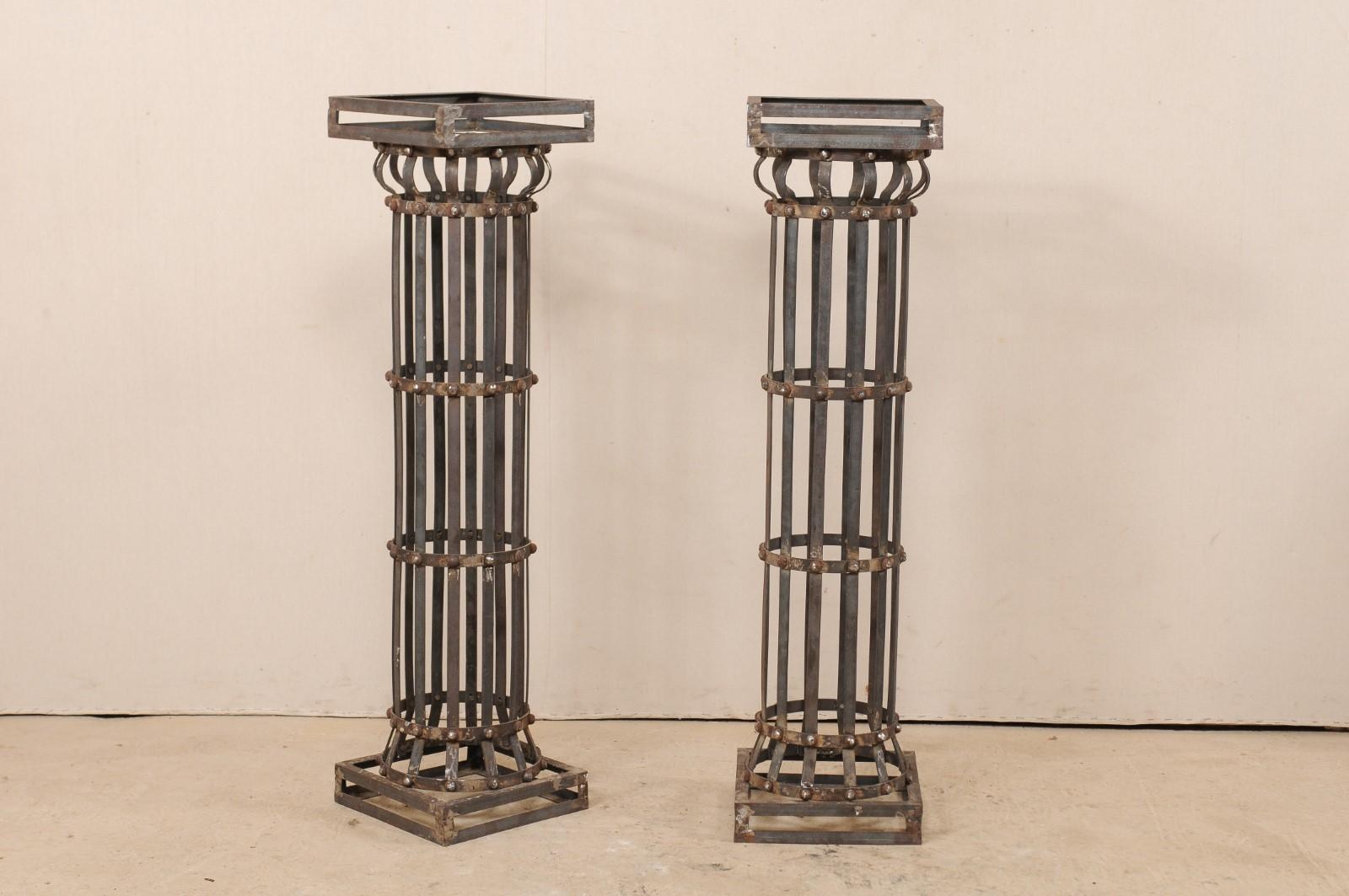 A pair of contemporary American iron architectural columns. This pair of vintage columns feature a round shaped shaft, comprised of vertically set iron strips, with a square-shaped capital and base, and stand at approximately 4.25 feet in height.