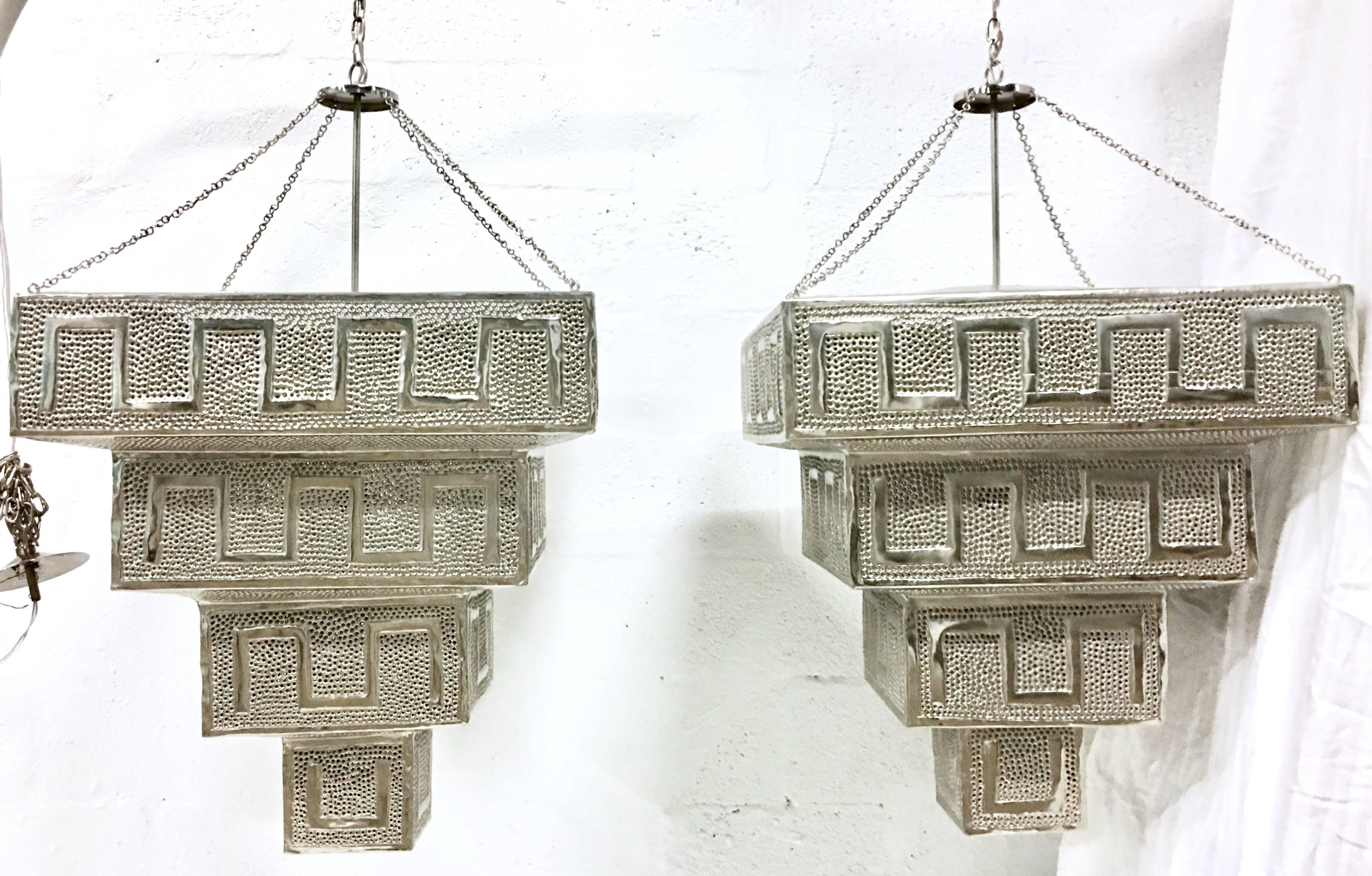 Contemporary pair of monumental silver steel and chrome pierced triple layer Moroccan style chandeliers. These Arabesque inspired works of art have a geometric raised Greek key pattern with perforated detail which allows the light to play of the