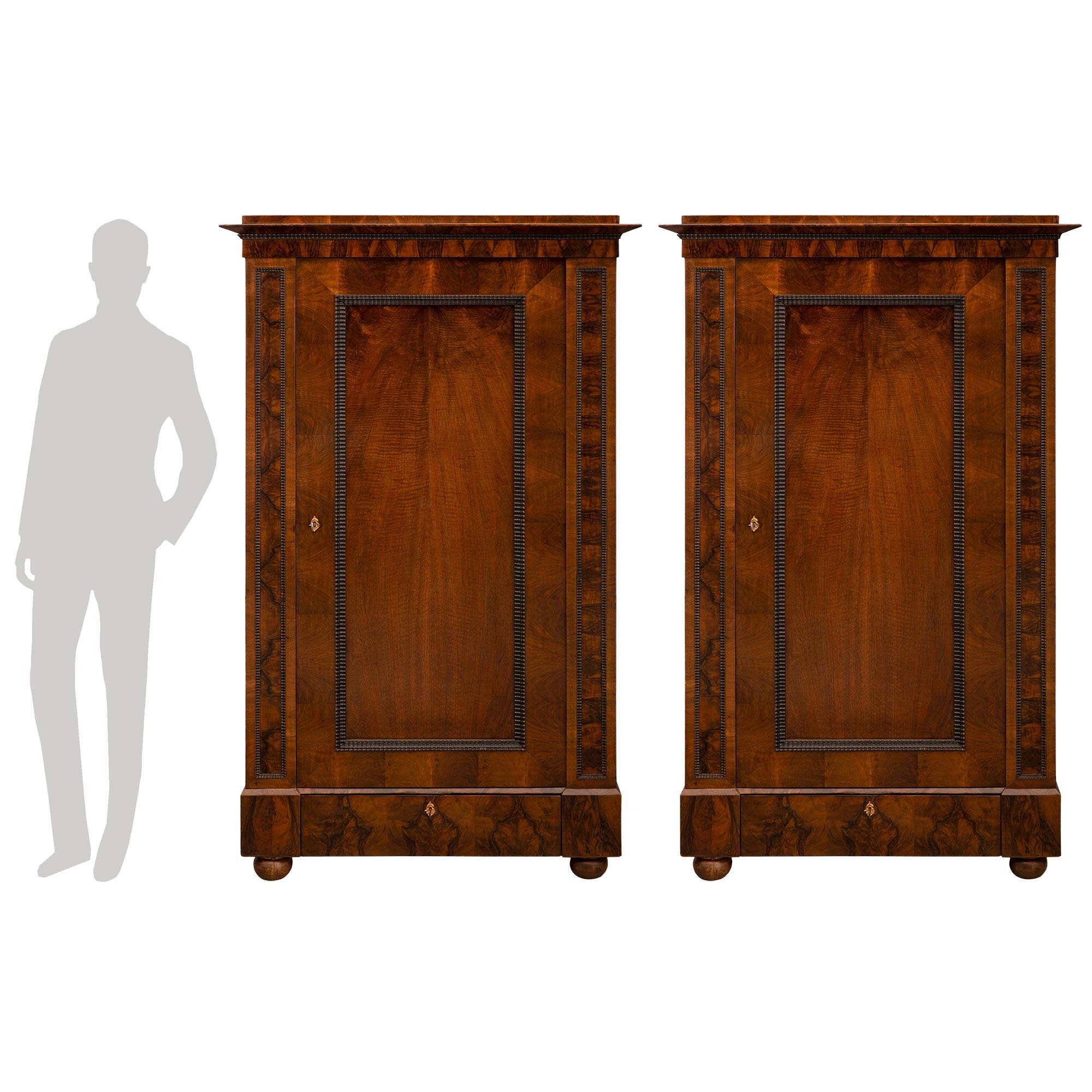 A striking and large scale pair of Continental early 19th century Neo-Classical st. Walnut and ebonized Fruitwood cabinets. Each cabinet is raised by four elegant walnut bun feet below a straight frieze with a single drawer decorated with an ormolu