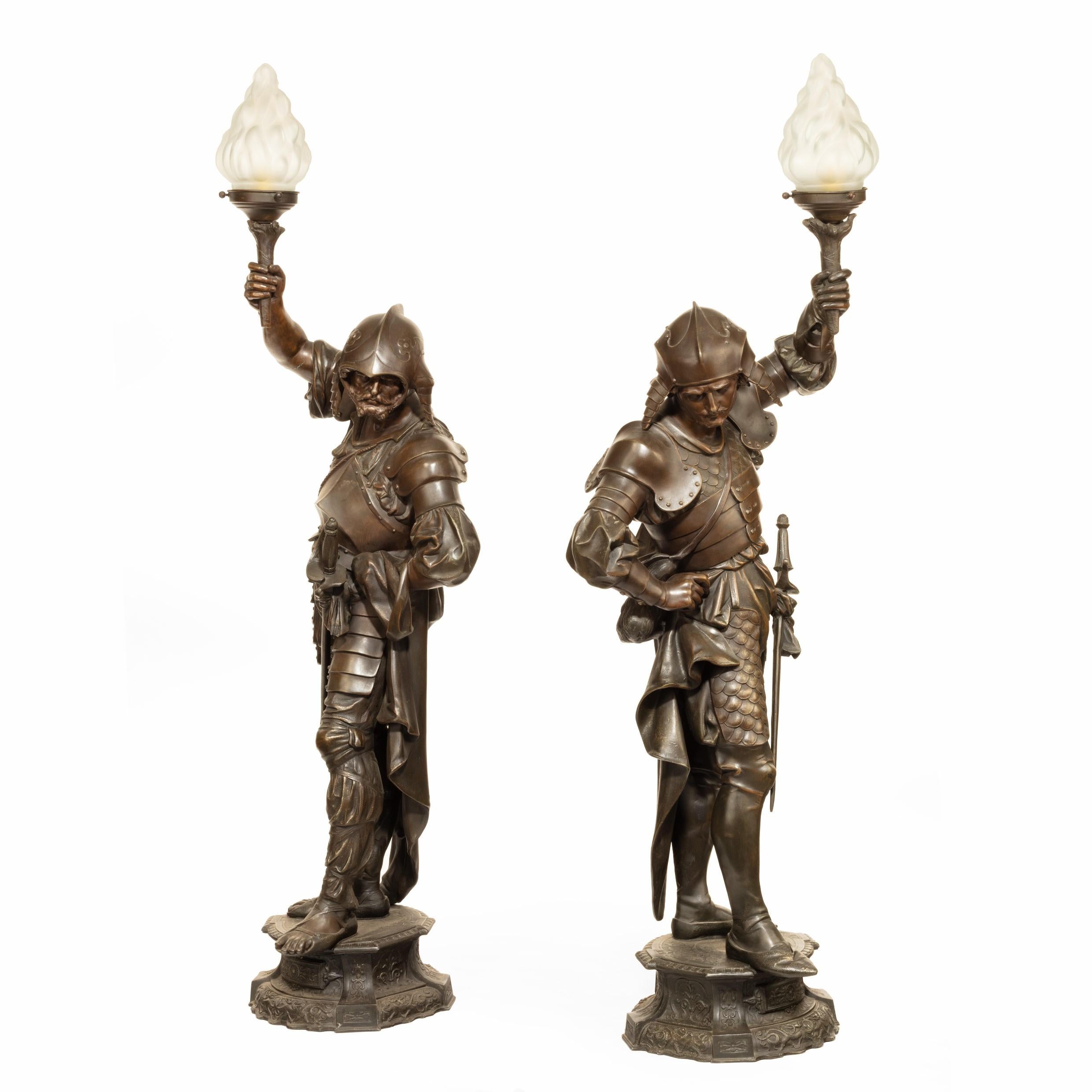 A pair of Continental bronzed-spelter gasoliers, each in the form of a soldier wearing heavy plate armor and ridged helmet with ear flaps, holding aloft a torch with a frosted glass flambé shade, on a flanged base, fitted for electricity, circa 1900.