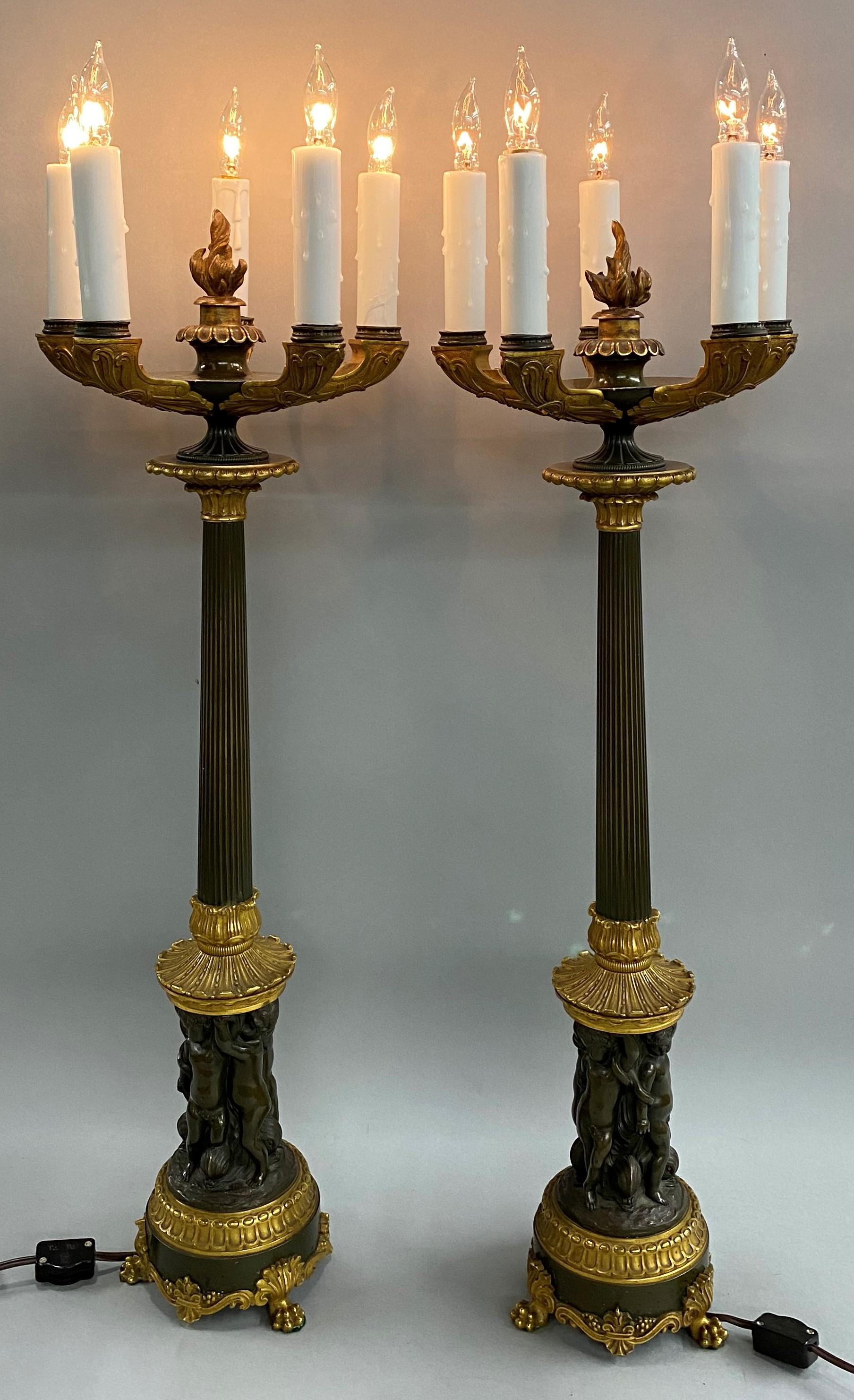 A fine quality pair of Continental gilt and patinated bronze electrified five-light candelabra lamps with center gilt flames, cast gilt scrollwork arms, reeded column stems, featuring multiple cast bronze putti surmounting a round bronze base with