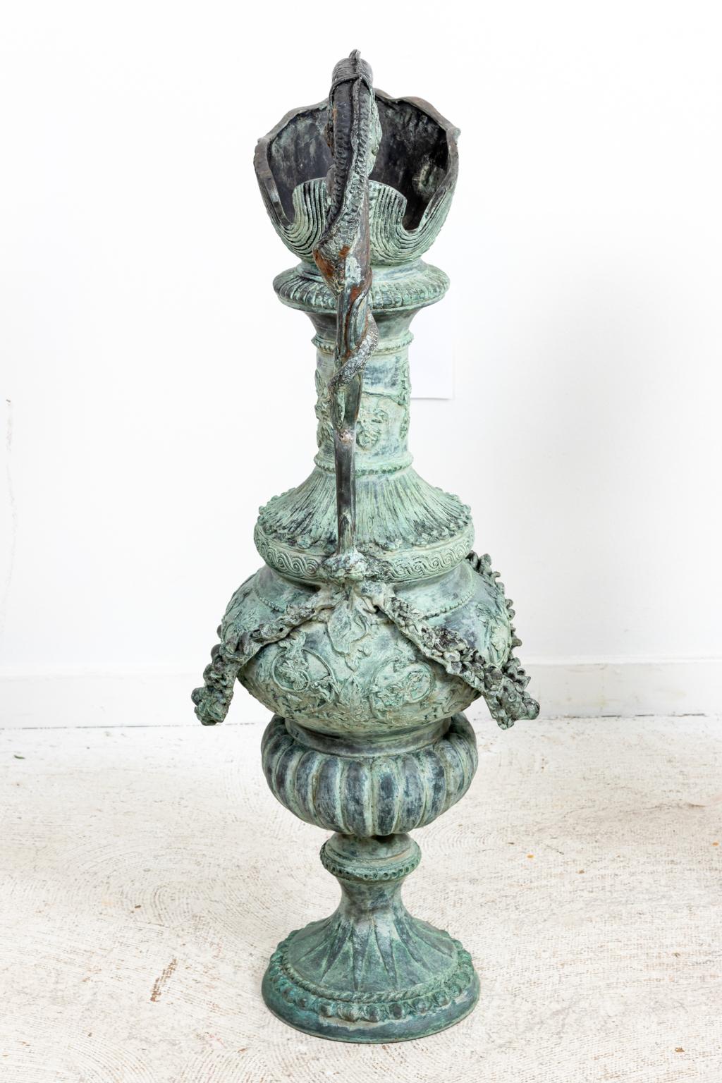 Pair of Continental style verdigris bronze urns with single handle and floral Roman swags. The piece is further detailed with motifs such as scrolled foliage,putti half figures, gadroon trim on the base, and scallop shell trim on the rim. Please