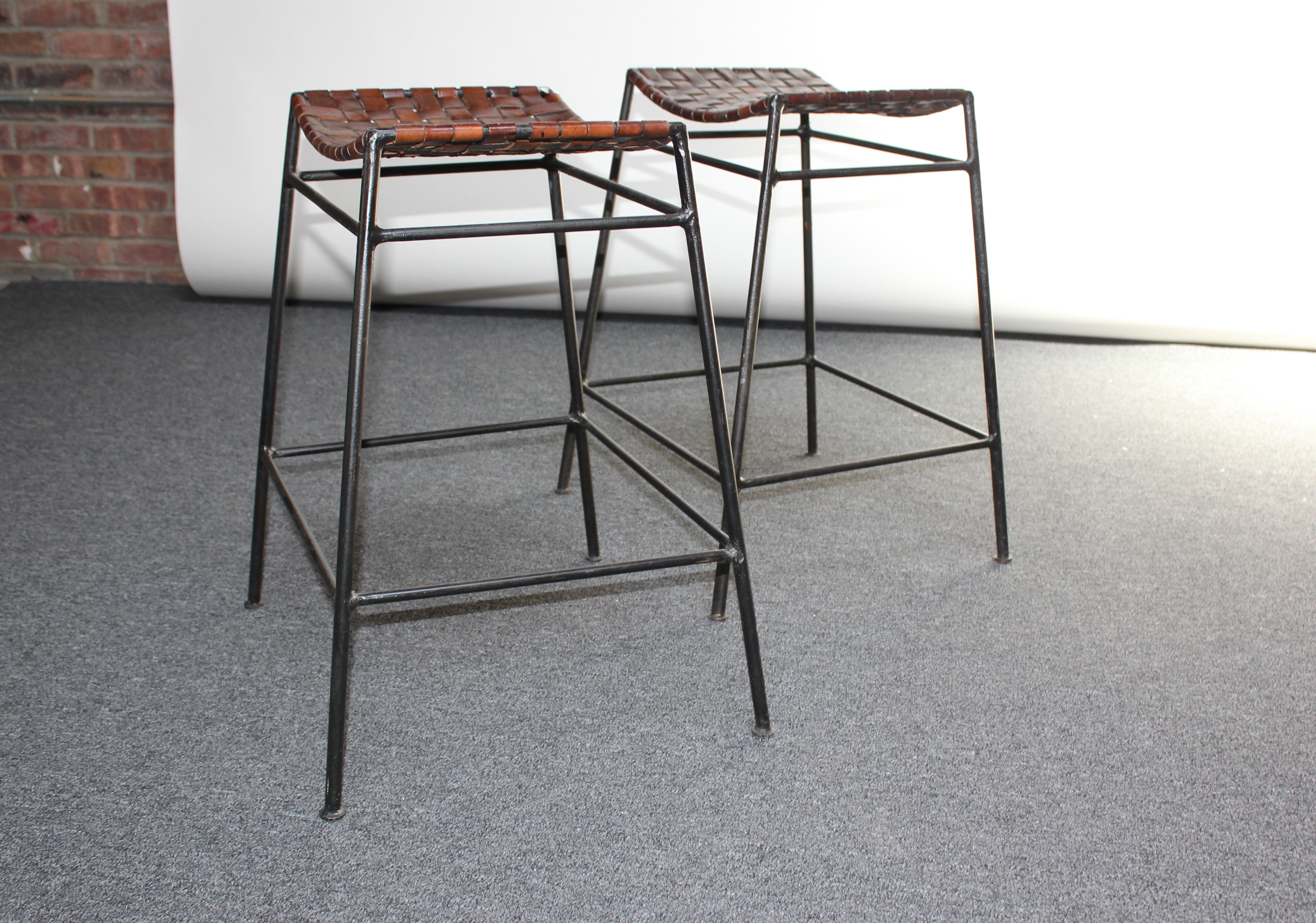 Pair of counter-height stools in iron and woven brown leather in the style of Swift and Monell (ca. 1980s, USA). Leather is richly patinated, showing wear from age and use, but sturdy and fully intact. Iron frames retain their original black paint,