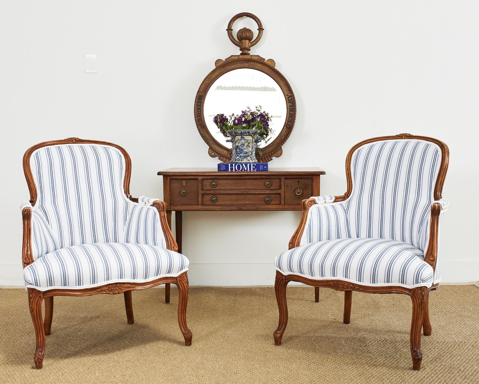 Charming pair of country French provincial style bergere armchairs featuring a blue and white fabric having a French ticking stripe style design. The molded frames feature scrolls and carved floral motifs with a gracefully curved back, arms, and