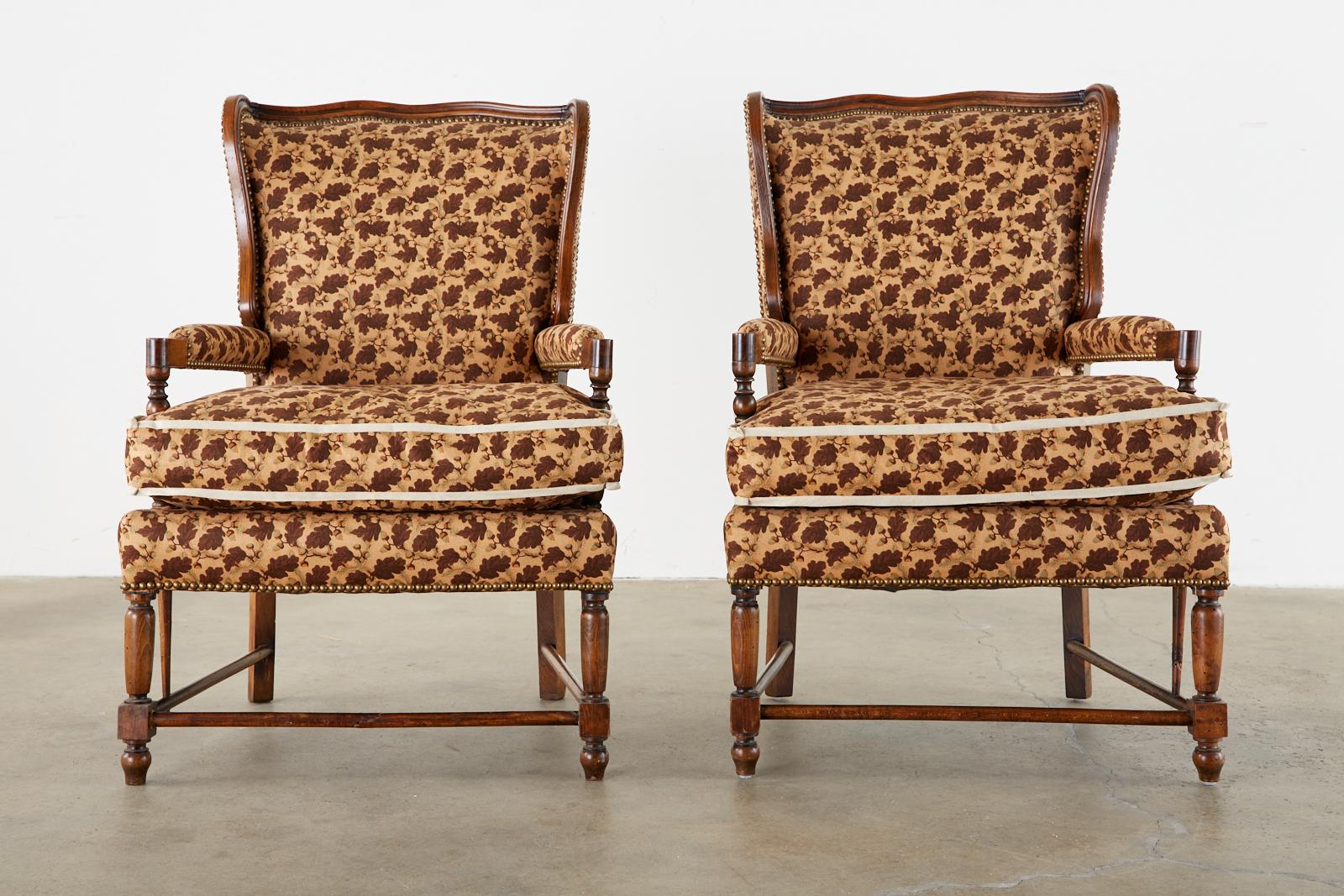 Distinctive pair of French wingback chairs or armchairs made in the country Provincial style. The pair feature a generous carved frame with a serpentine crest rail and wings. Upholstered with a foliate and acorn motif fabric fitted with a deep loose