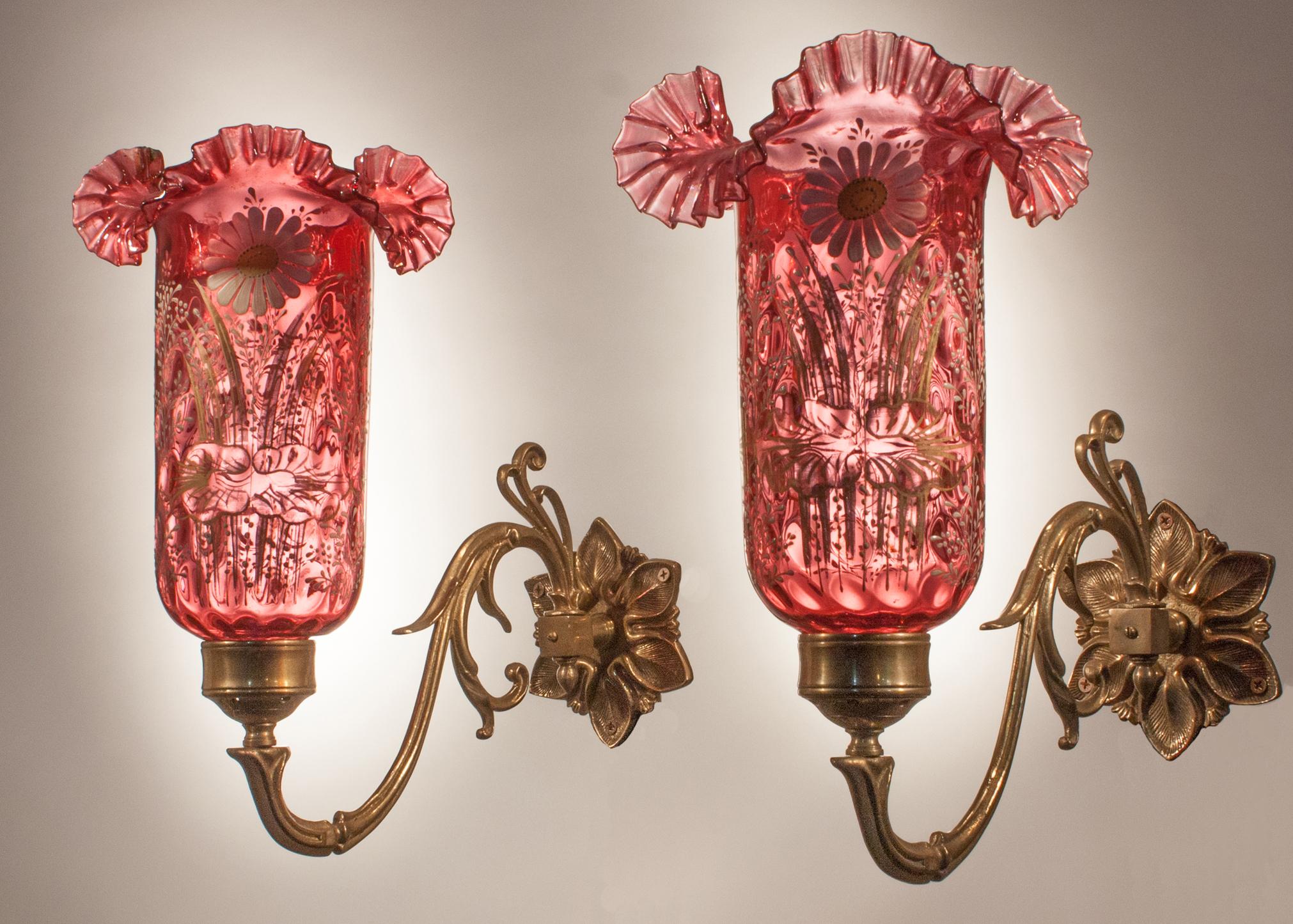An unusual pair of cranberry colored hand blown glass sconce shades hand painted with a floral relief motif. These circa 1920 Anglo-Indian shades feature delicate ruffling on the rims, with circular dimpling from top to bottom. The wall sconces take