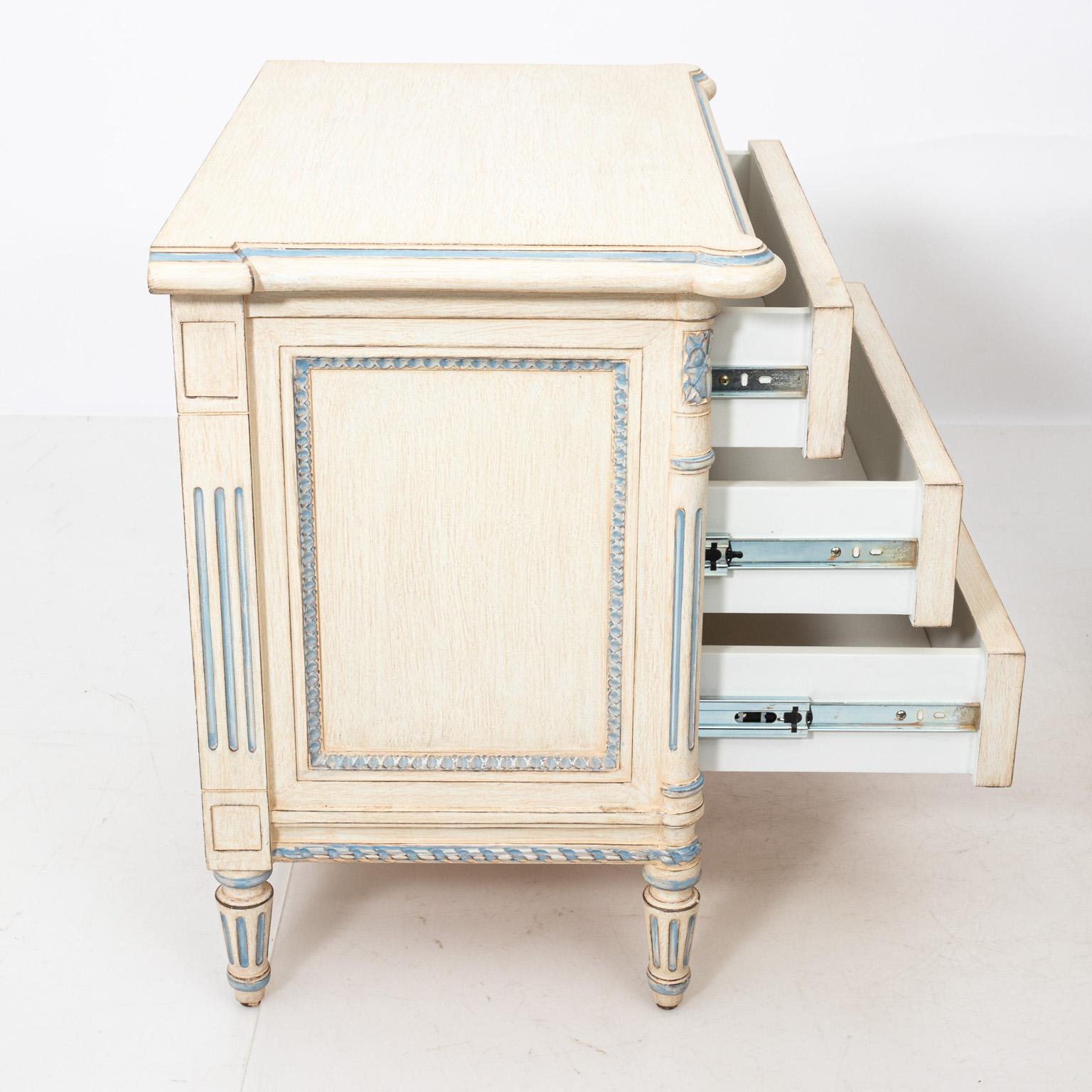 Louis XV style pair of cream painted bedside tables or commodes with three drawers and metal hardware in the manner of Julia Gray, circa 1980s. They also feature blue accent trim and carved rosettes. Please note of wear consistent with age. Made in