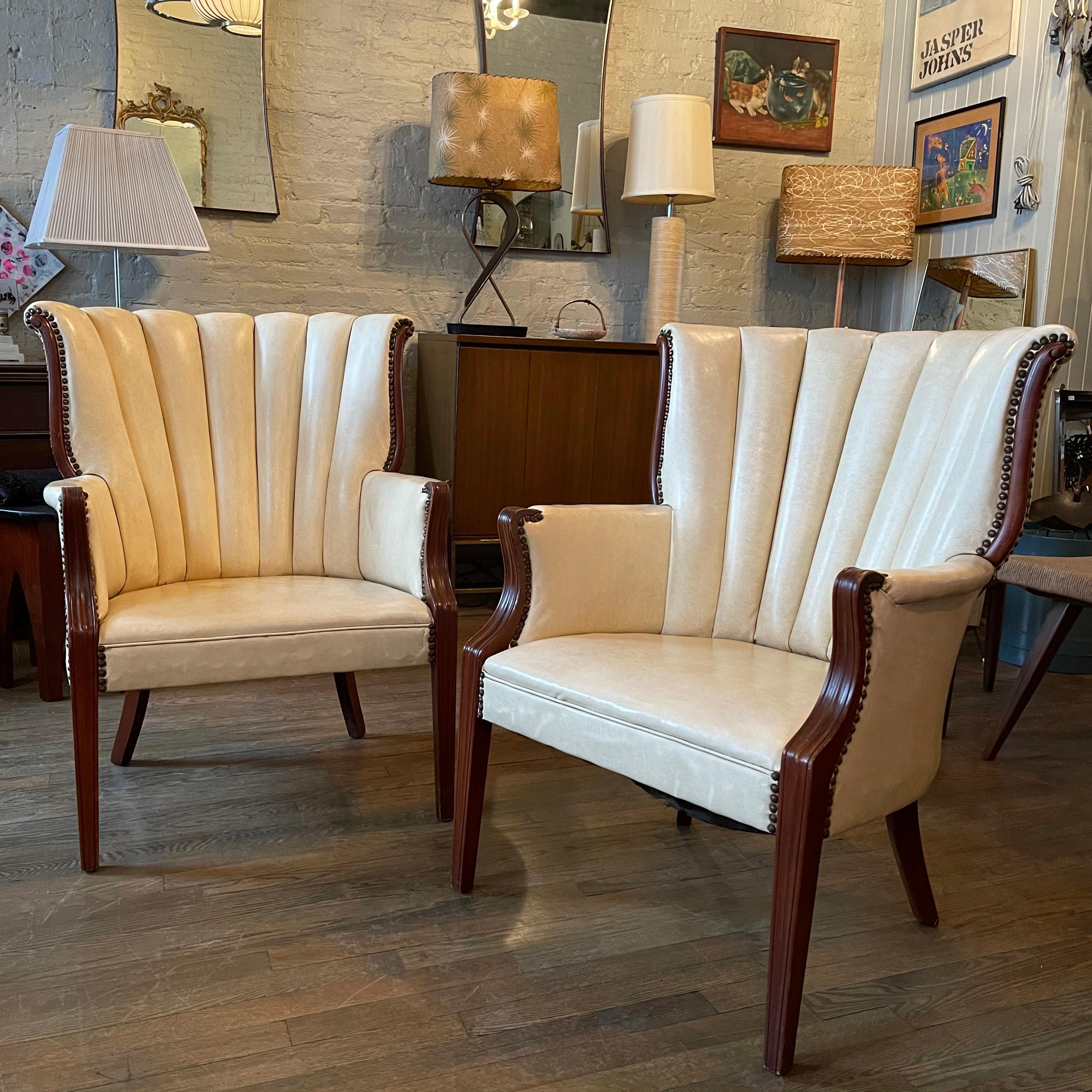 Pair of midcentury, Regency style, mahogany wingback armchairs feature cream vinyl upholstery with scalloped back and studded nailhead detail. The arm height is 23.5 inches.
