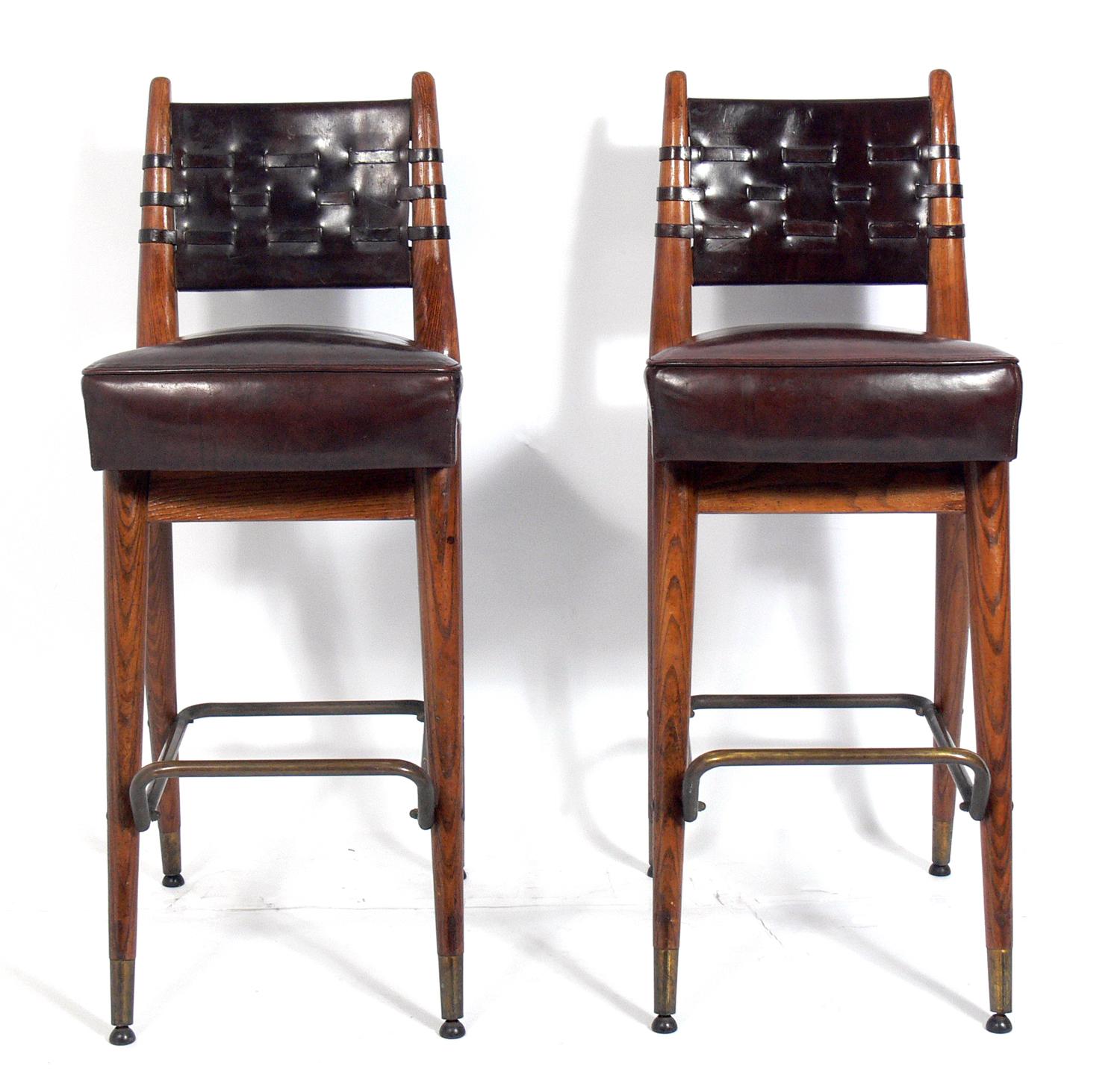 Pair of curvaceous oak leather and brass bar stools, American, circa 1960s. They retain their original deep brown leather upholstery with interesting woven leather backs. Curvaceous oak frames with brass foot rests and sabots. They retain their warm