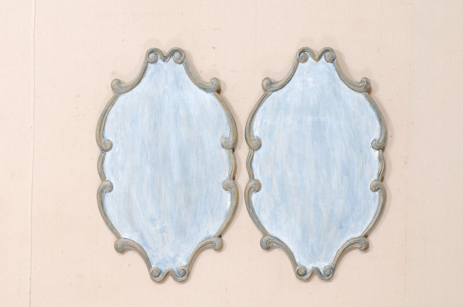 A pair of Italian style hand carved and painted wood hanging wall plaques. This pair of wall mounted wood plaques feature hand carved, mostly oval-shaped bodies, with a lovely boarder frame comprised of delicate carved loose scrolls. The wood has a