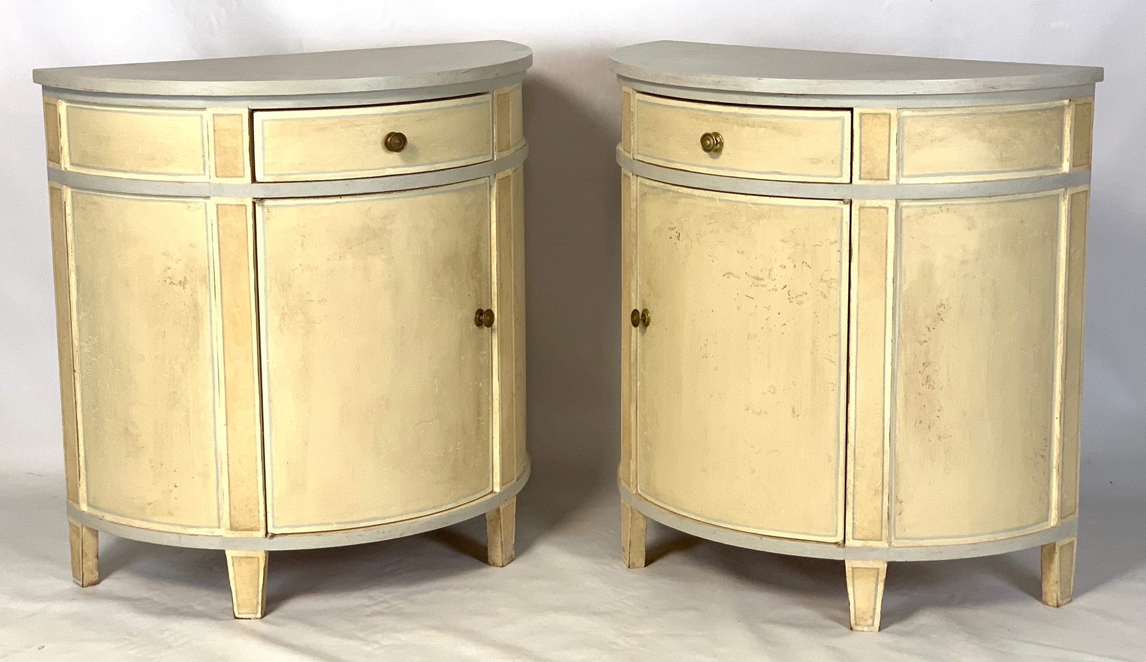 A pair of mid-20th century custom made and paint decorated bedside tables in a palette of distressed cream and pale gray each having a single drawer over opposing cabinet doors revealing two shelves.