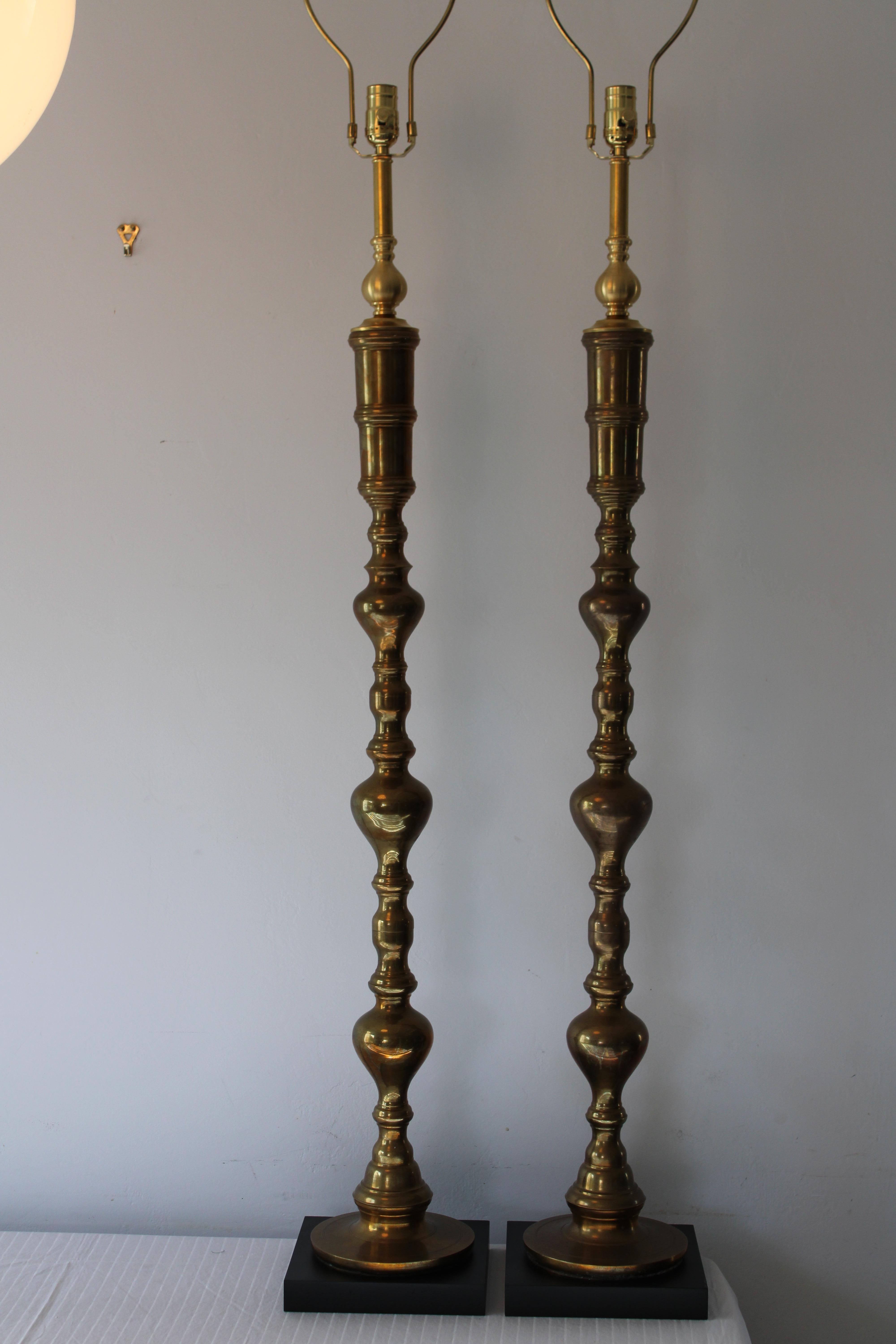 A matched pair of tall solid brass lamps with black painted wood bases. We took a pair of brass candlesticks and modified them to produce an elegant pair of brass lamps. Lamps measure 46.5