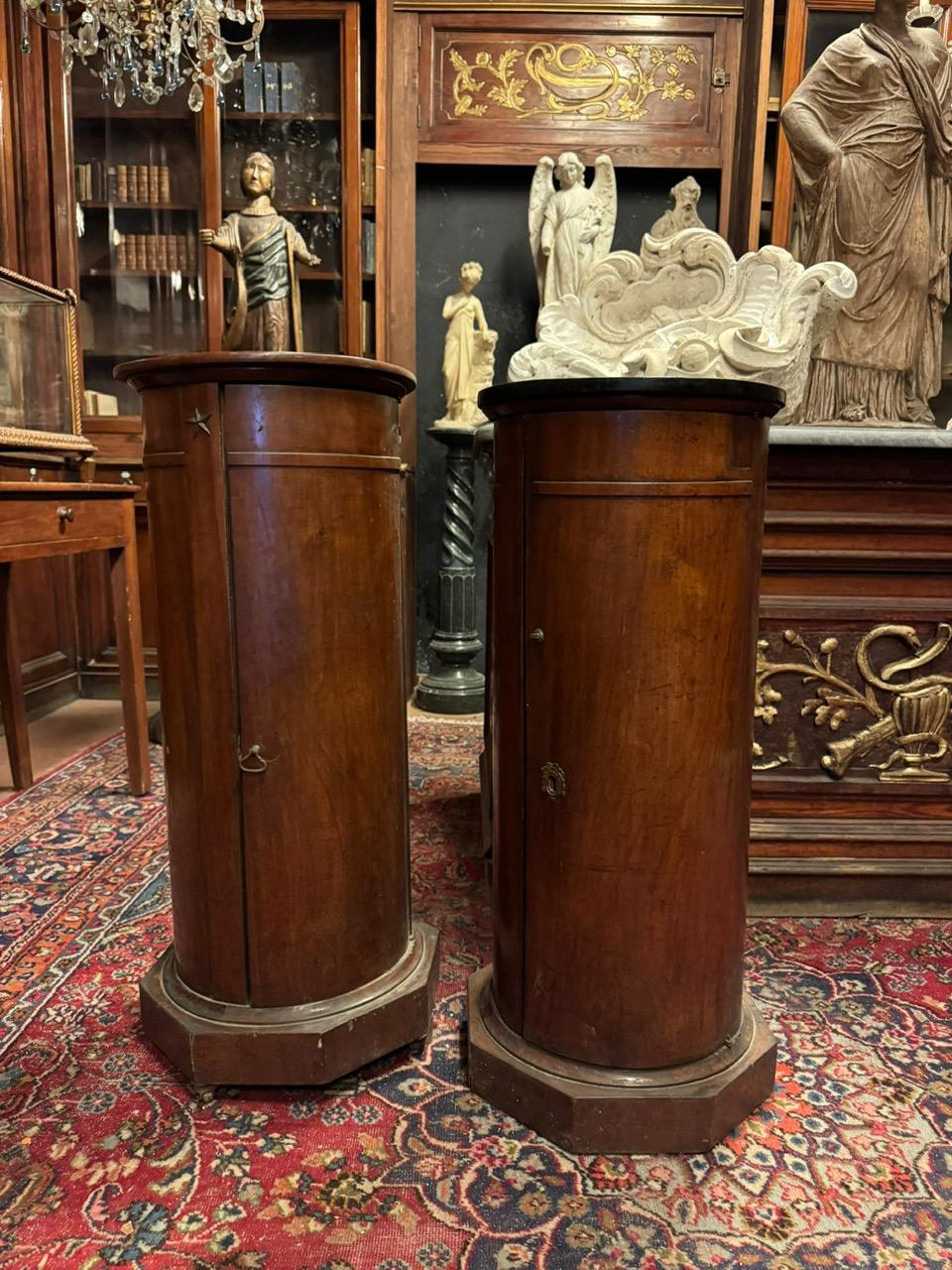 A pair of antique walnut bedside tables. They feature a cylindrical structure with an octagonal base.
They have a glossy finish that highlights the wood grain. They feature a vertical door with internal storage shelves.
The shape of the bedside