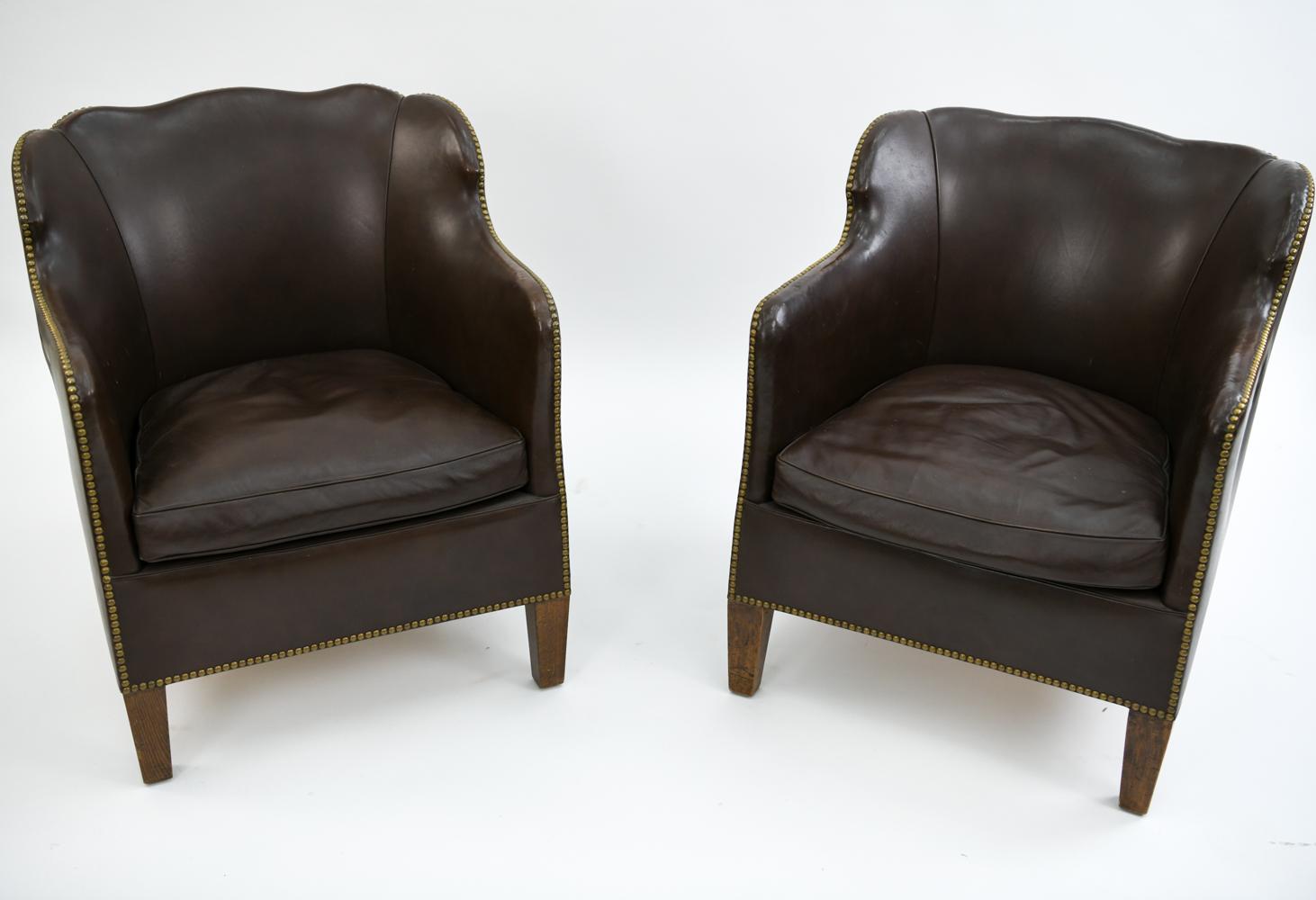 This is a handsome pair of Danish library chairs. Upholstered in leather, these chairs feature brass tacked edges and scalloped backrests. These chairs would be attractive in both modern or traditional interiors for their timeless appearance.