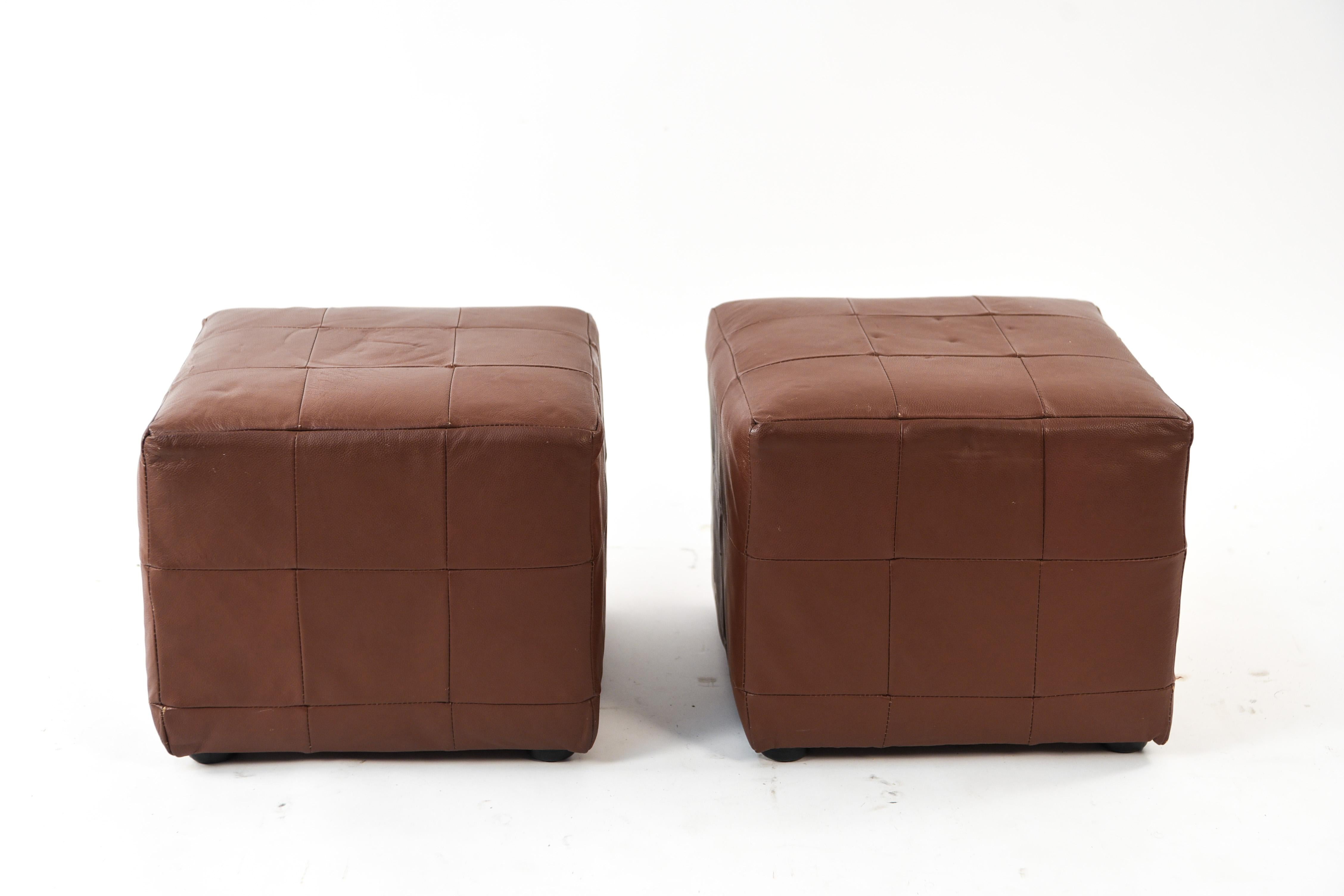 This pair of midcentury ottomans are upholstered in leather. These ottomans are in a simple cube shape. The upholstery mirrors the cube shape by creating visual squares over the surface.