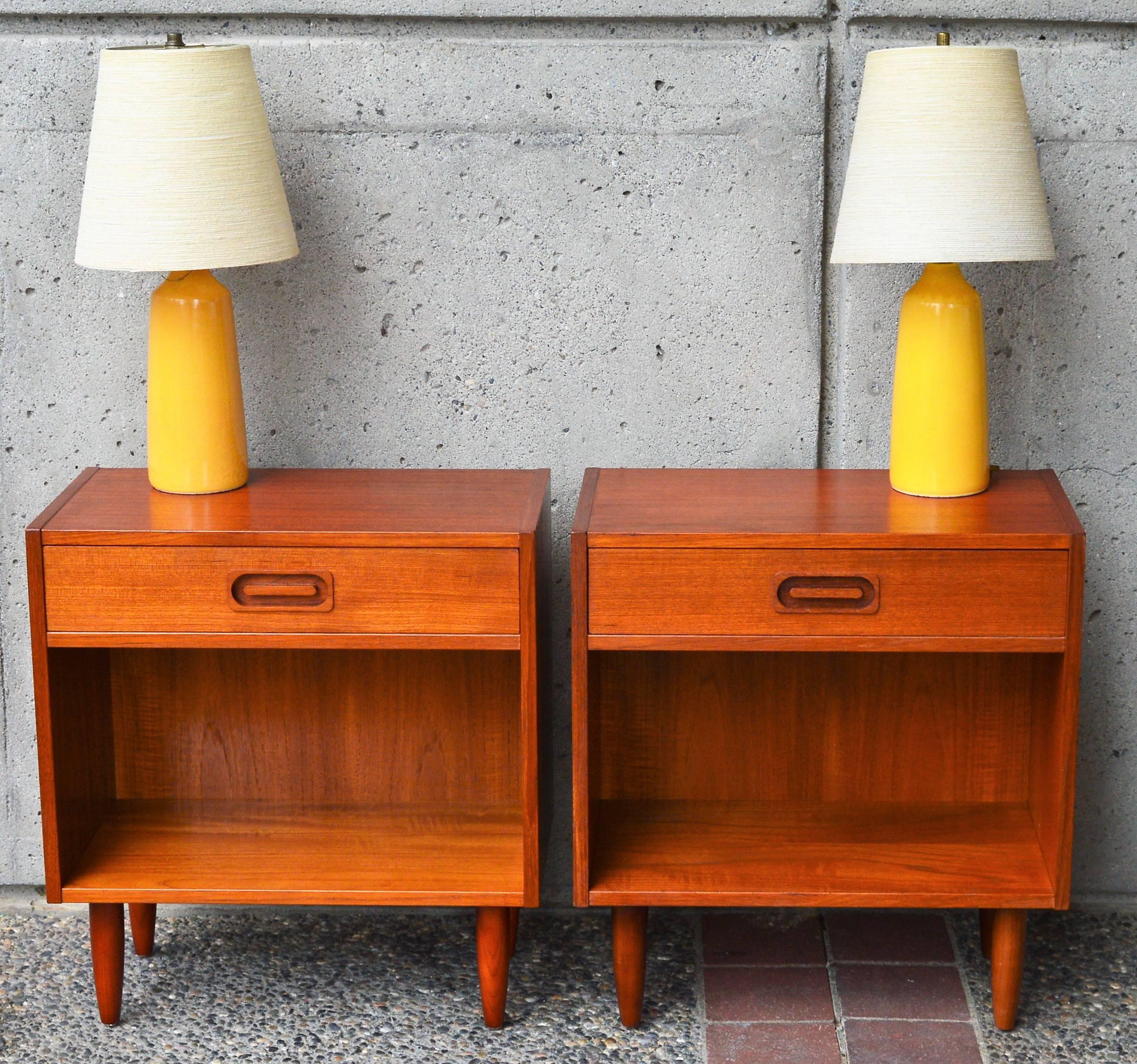 This fabulous pair of Danish modern teak bedside tables or nightstands feature a cubic design with one drawer with wood frame construction and dovetail joints as well as an open lower cabinet. Finished with tapering conical legs and sweet oval inset