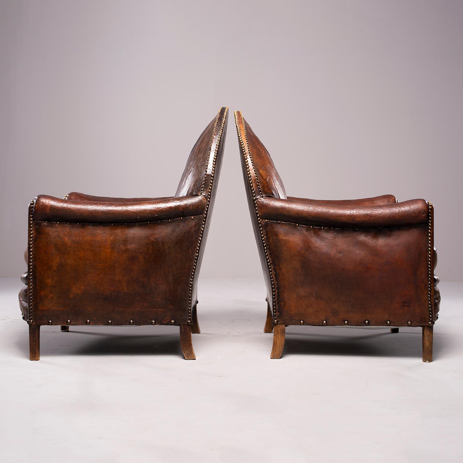 Pair of classic French Art Deco brown leather club chairs in original condition. Camel back seat rests, rolled arms, brass nail head trim. Leather is in original condition and has visible wear throughout, see detail photos. Arms are 24” high. Seats