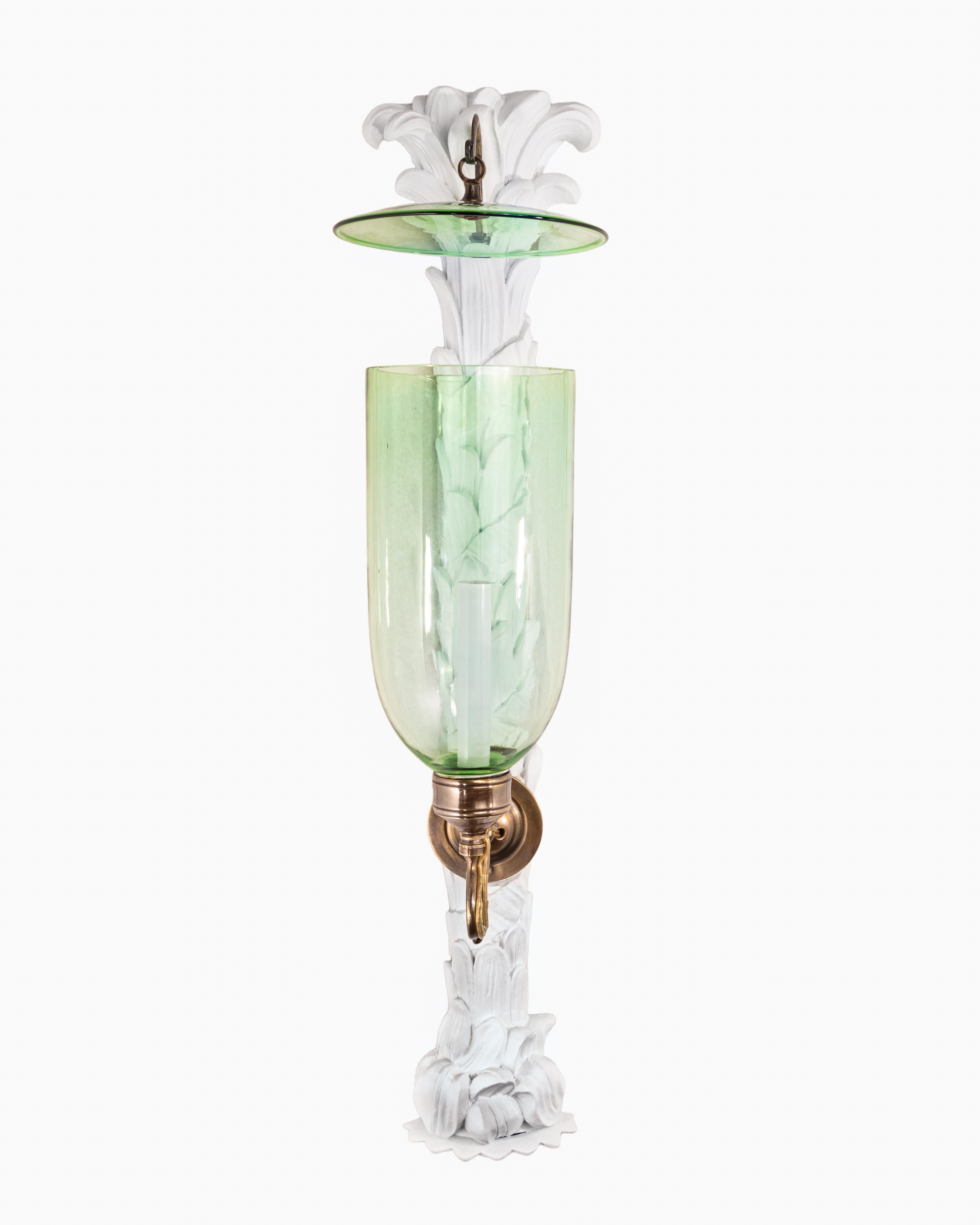 A pair of hand-carved palm branch styled mahogany backplates painted in a plaster finish with patinated brass sconce fittings and green glass hurricane shades. Electrified with one candelabra base socket per sconce.

Our custom hurricane sconces are