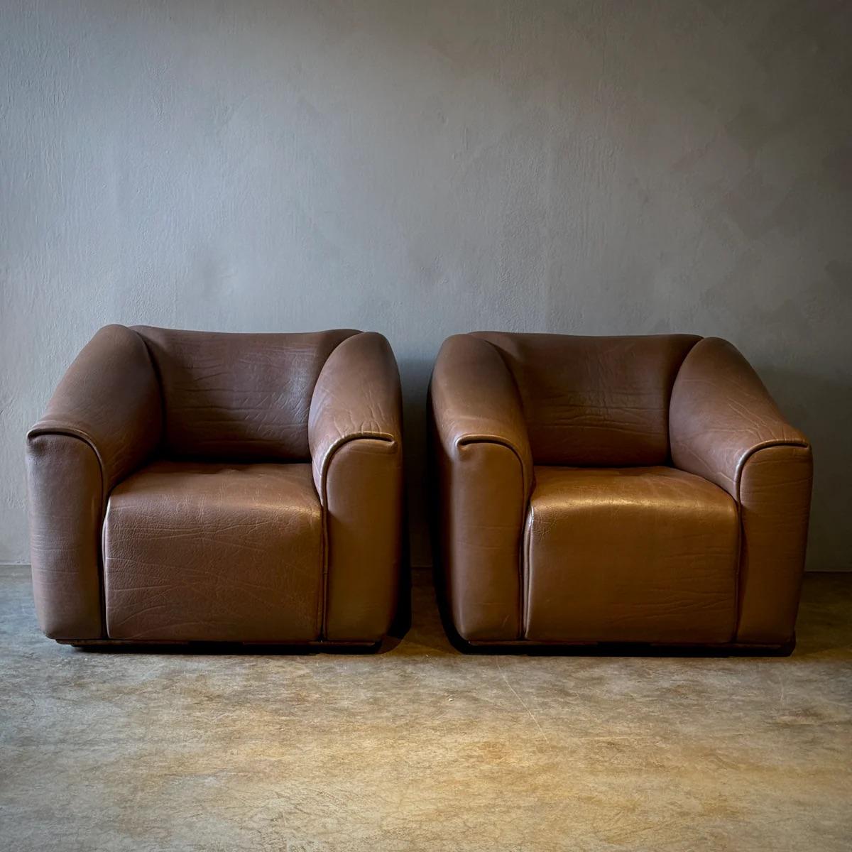 Dieses Set besteht aus zwei robusten de Sede Lounge-Sesseln und einer passenden Ottomane, die alle mit prächtigem Leder gepolstert sind, das anmutig gealtert ist und eine warme, einladende Patina angenommen hat. Die plüschigen Kissen und die