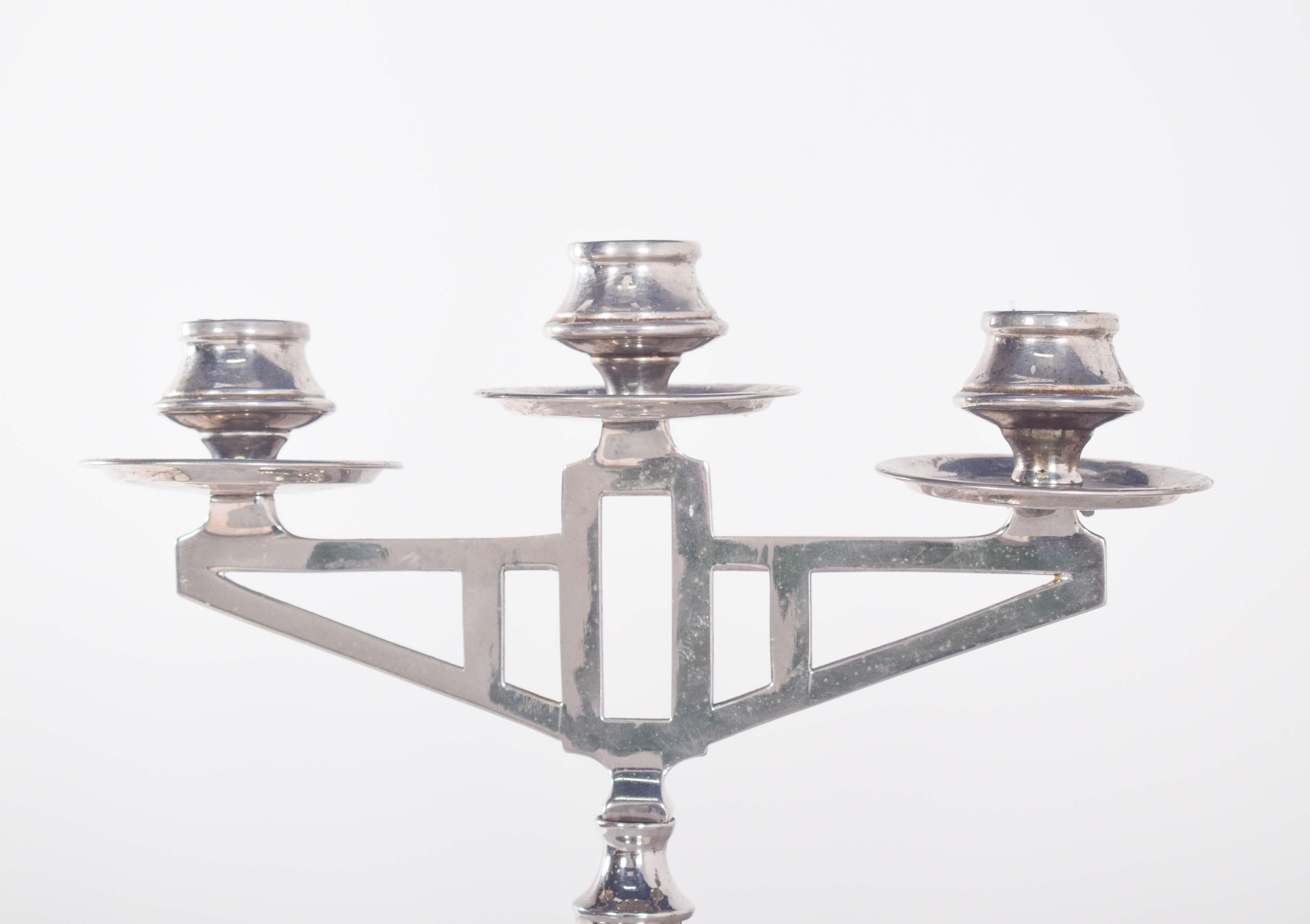 Paar Deco-Kerzenständer aus silbernem Metall. Mit drei symmetrisch angeordneten Kerzenarmen, die ein ausgewogenes und harmonisches Bild ergeben. Die zentrale Struktur besteht aus durchbrochenen rechteckigen und dreieckigen Elementen, die einen