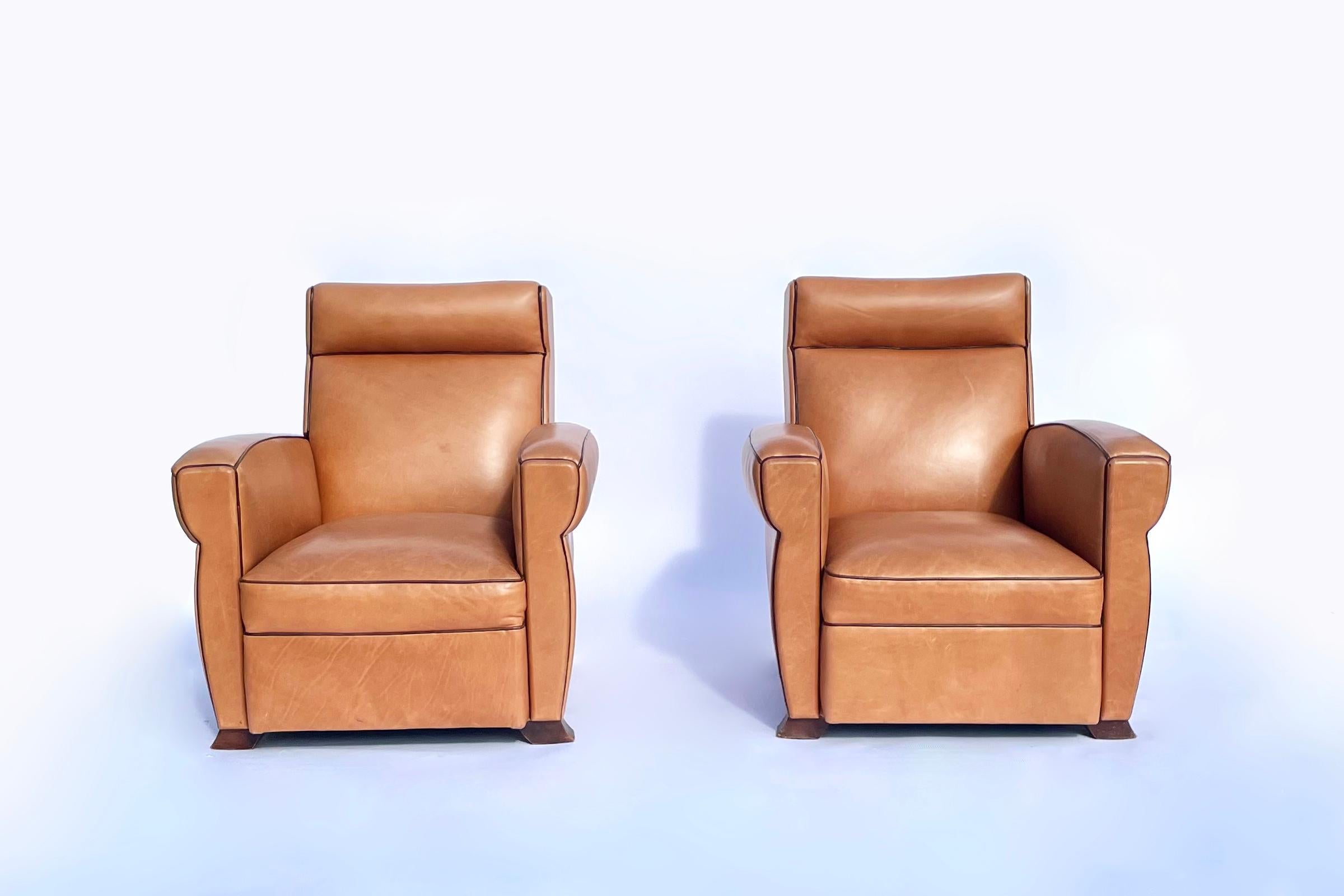 Zwei sehr bequeme Art-Déco-Clubsessel, gepolstert mit kamelhaarfarbenem Leder, mit braunen Ledereinfassungen und patinierten Messingnagelköpfen. Sie wurden etwa 2007 wie abgebildet neu gepolstert. Original Mahagonifüße im Originalzustand. 