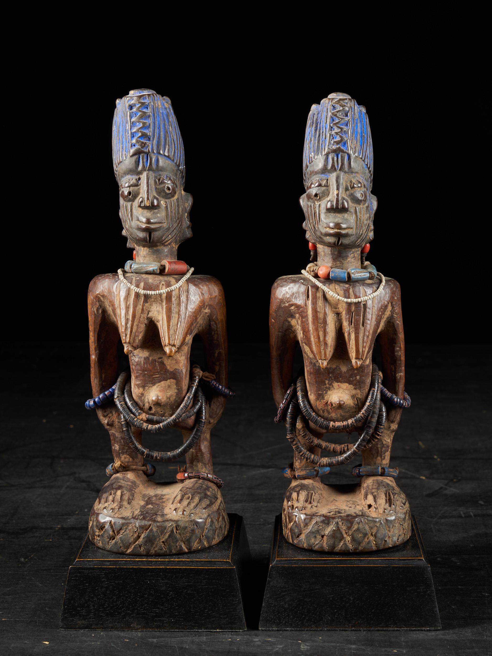 A pair of female ‘ibeji’ twin figures of the Yoruba people. From south-west Nigeria . Both ‘ibeji figures’ wear a high plaited coiffure, coloured blue by means of ‘bluing’ (‘Reckitt’s Blue’). Both bodies and the round plinth show remains of