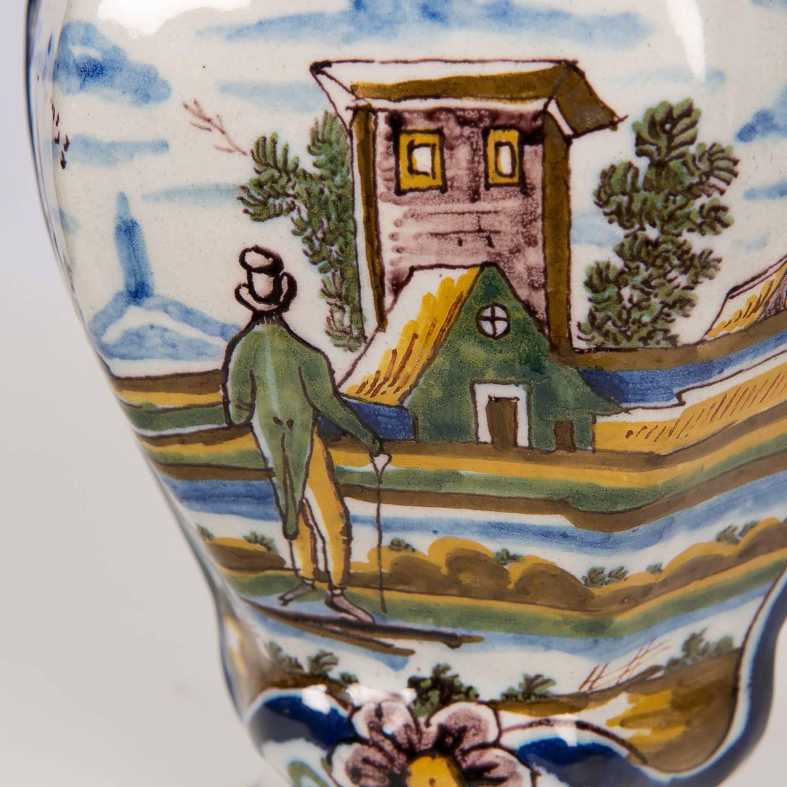 We are pleased to offer this lovely pair of Dutch Delft mantle vases showing a gentleman with a walking stick looking out across a river. 
On the other bank we see colorful houses and a church. The scene is outlined with a traditional dark blue line