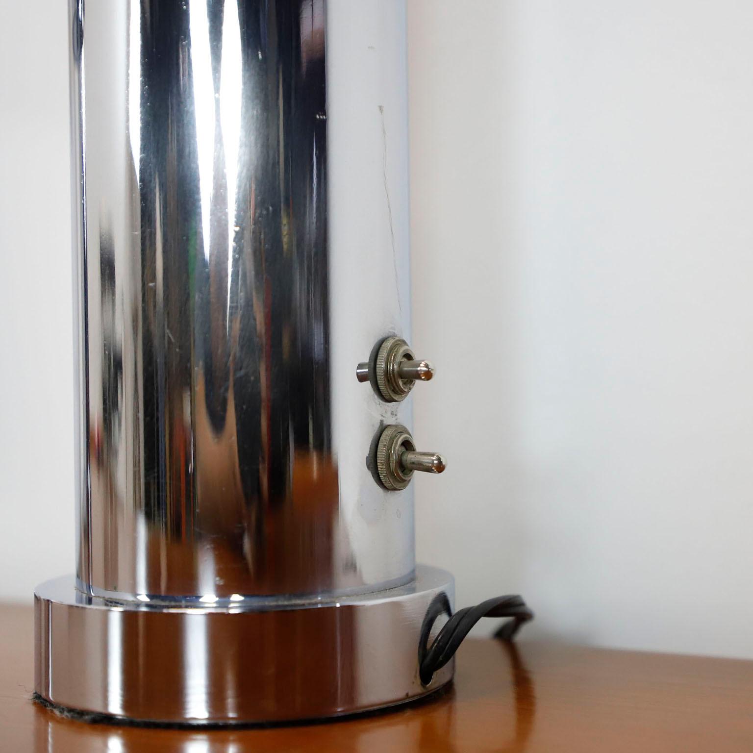 Interessante coppia di lampade da tavolo cromate, disegnate da Angelo Lelli per Lightolier, circa anni '60. Il corpo cilindrico della lampada è diviso in due in diagonale, con una sorgente luminosa nella sezione inferiore diretta verso l'alto e una